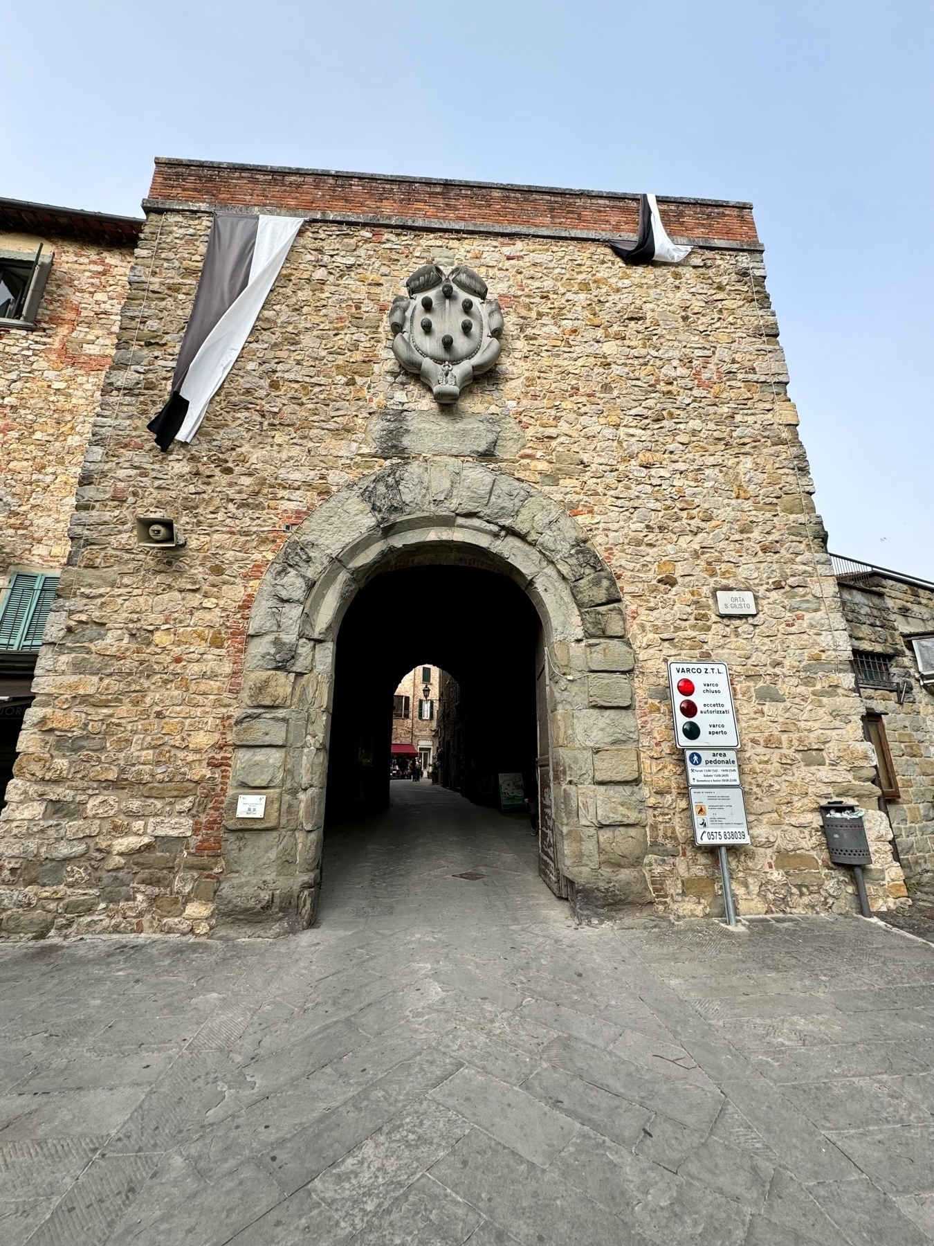A historical stone archway with a medieval crest above it, situated in a rustic town. Black, white, and grey flags are draped over the wall.