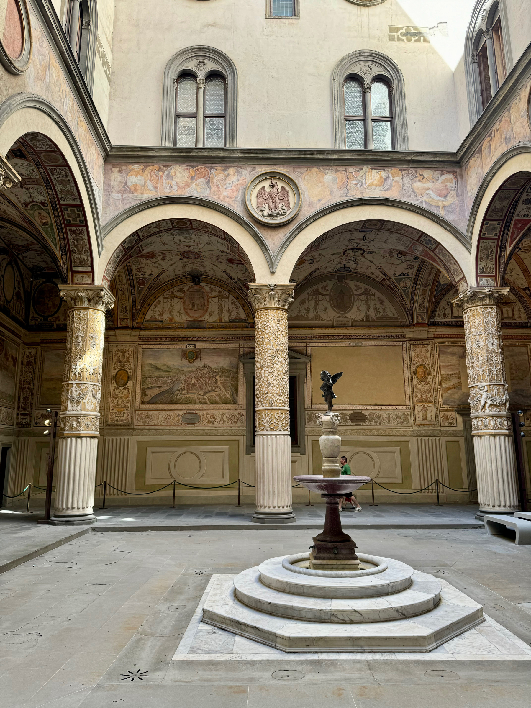 This image shows an interior courtyard featuring ornate columns, arched ceilings with detailed frescoes, and upper windows with stone frames. In the center, there is a fountain with a small statue on top. The courtyard is decorated with historical and artistic elements. 
