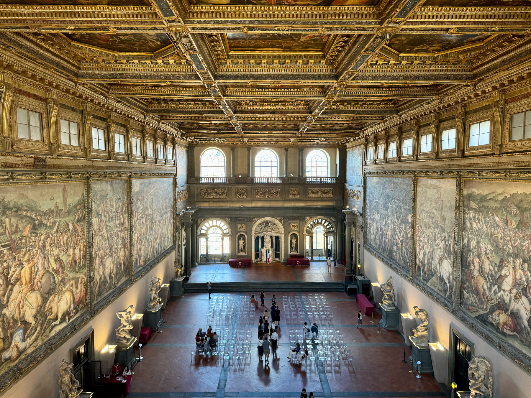 A grand hall with an ornate, gilded ceiling and large, detailed frescoes depicting historical battle scenes on the walls. Statues line the perimeter of the hall below the frescoes. The hall features tall arched windows and a few visitors walking. 
