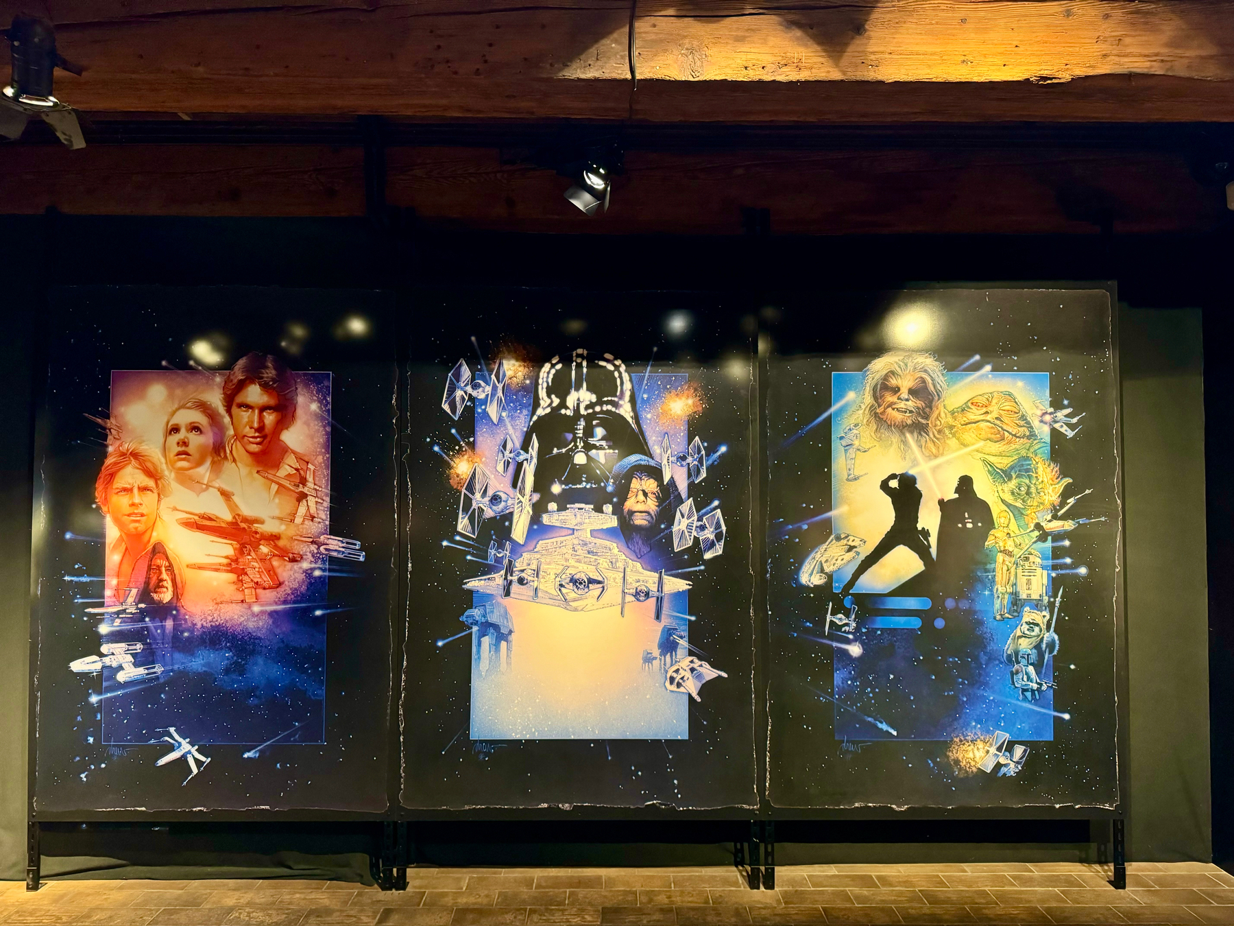 Three large posters depicting characters and spacecraft from the Star Wars franchise displayed on a black wall beneath wooden beams, with spotlights shining on them.