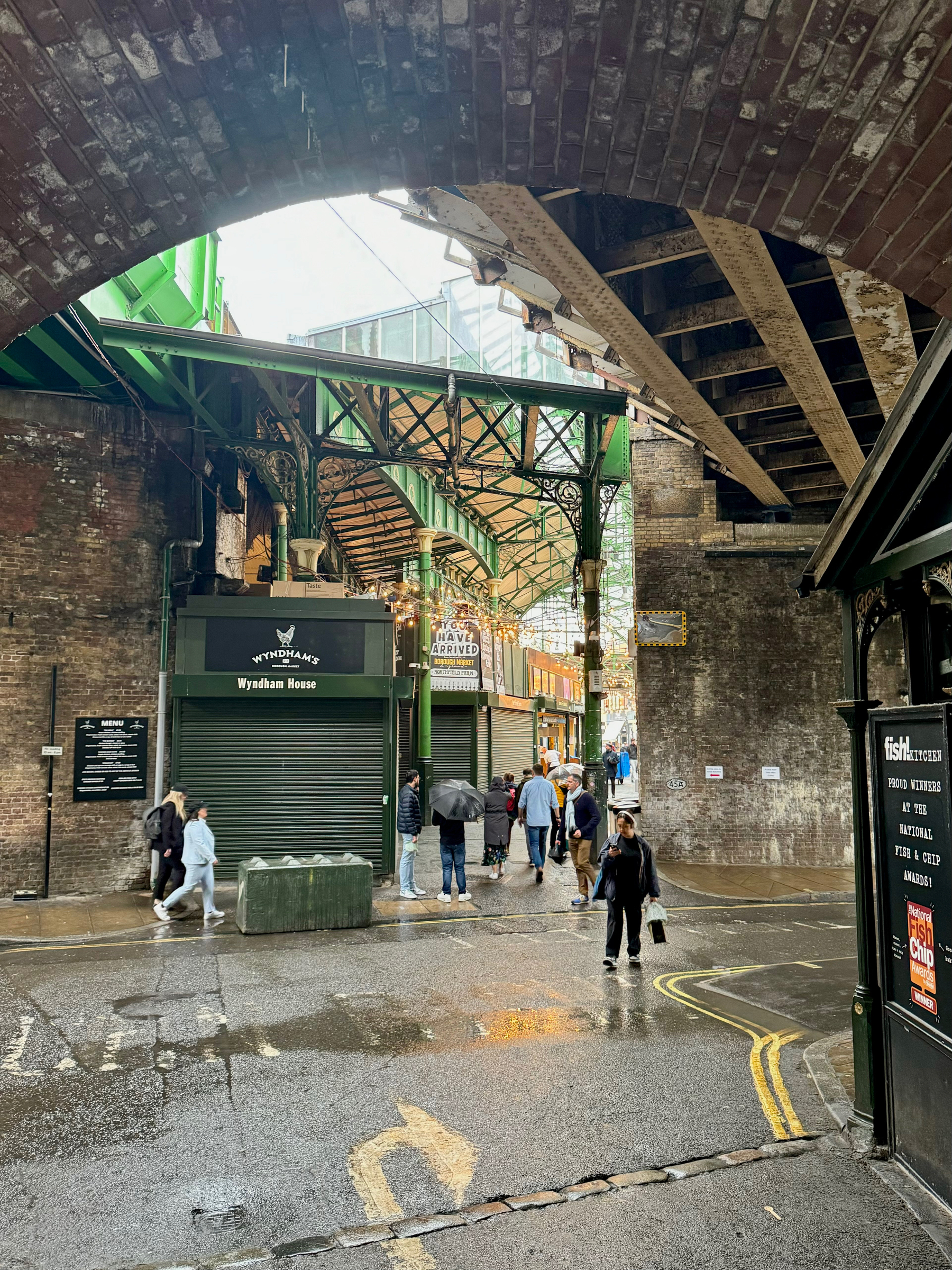 An urban street scene under a railway bridge with pedestrians walking and closed shopfronts, showcasing the architecture and atmosphere of a city on a damp, overcast day.