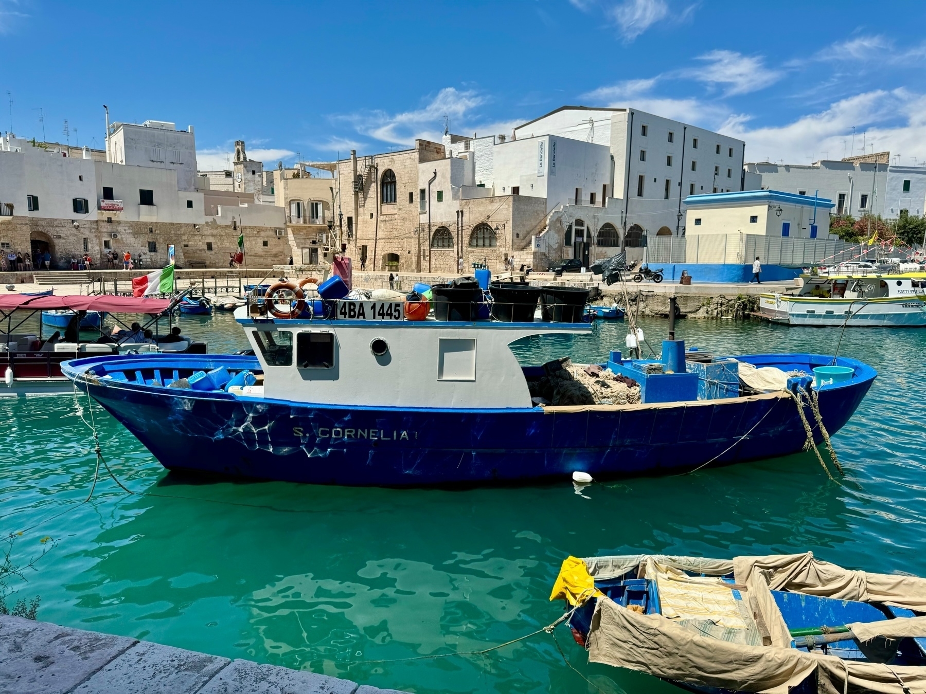 A blue fishing boat named “S. Cornelia” docked at a waterfront with several other boats nearby. The boat is moored in clear turquoise water against a backdrop of historic buildings with a mix of white and stone facades. 