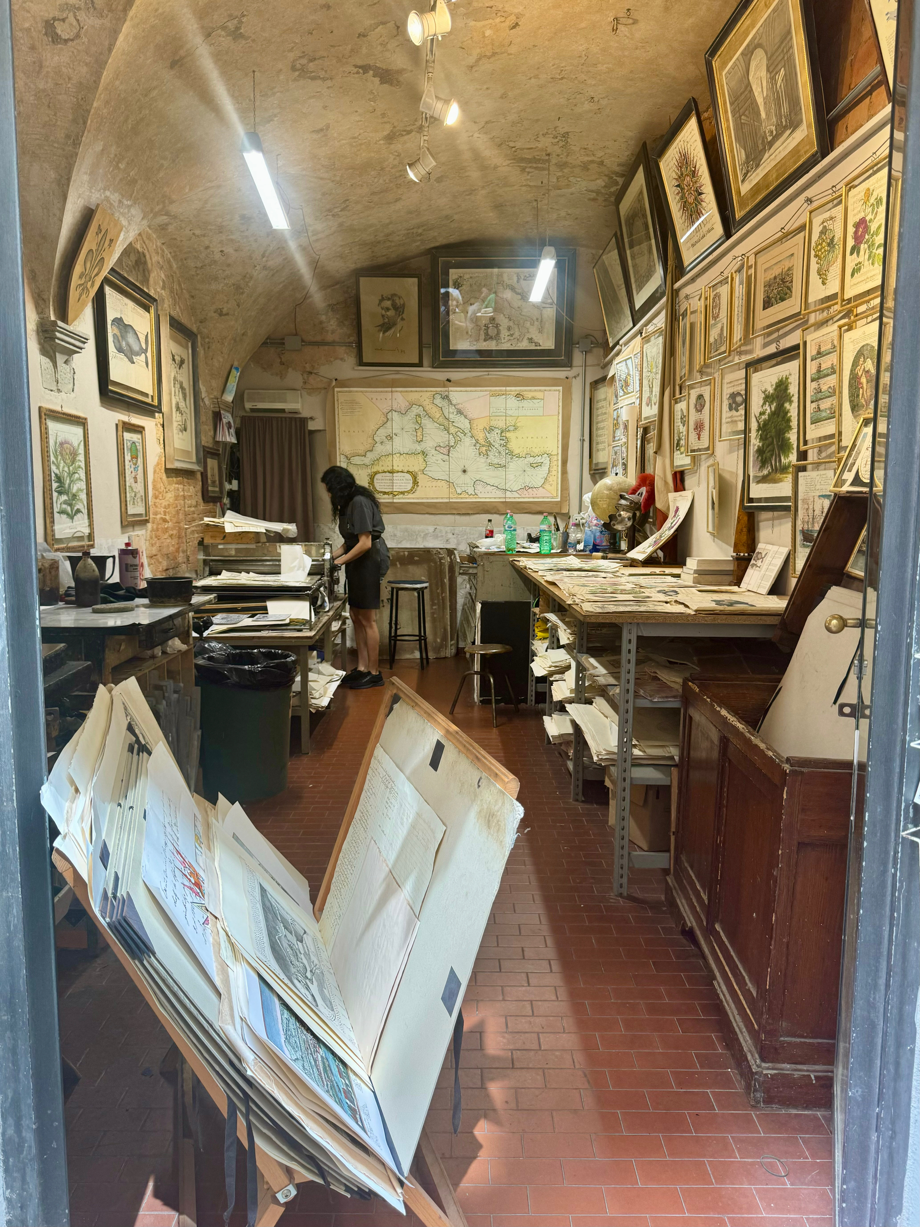 A small, cozy art studio with arched ceilings and exposed brick walls. The walls are adorned with various framed artworks, including botanical prints, portraits, and maps. A person is working at a table toward the back of the room. 