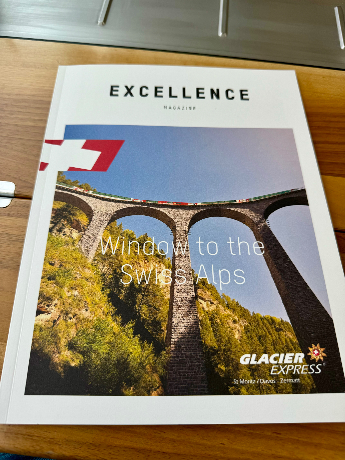 A magazine cover titled “EXCELLENCE MAGAZINE” featuring a photograph of a stone bridge with a train crossing, with the caption “Window to the Swiss Alps” and the Glacier Express logo, indicating routes from St. Moritz/Davos - Zermatt.