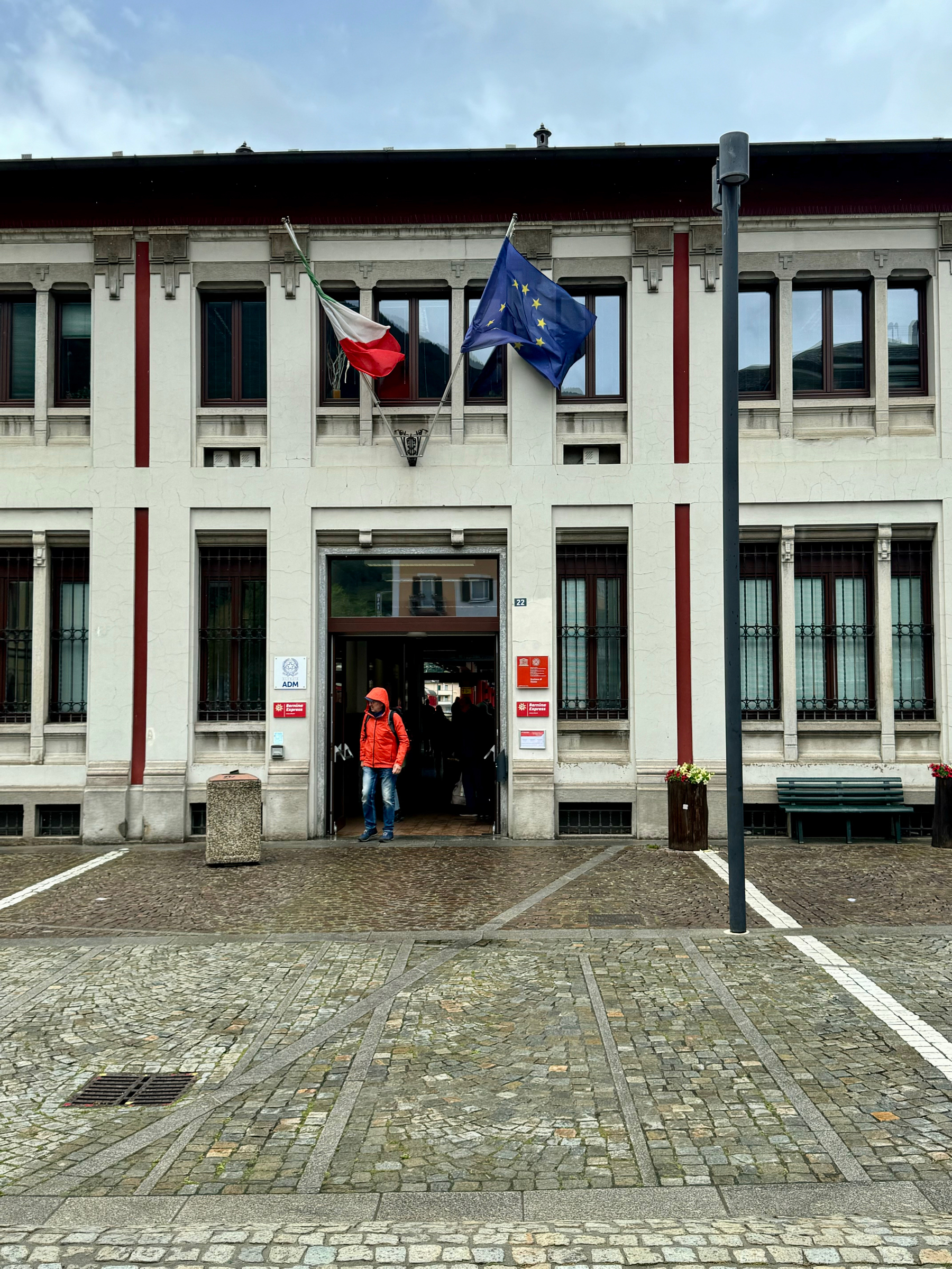 A person in an orange jacket standing at the entrance of a building adorned with Italian and European Union flags. The building has a white facade with red vertical accents and multiple windows. The ground is paved with cobblestones.