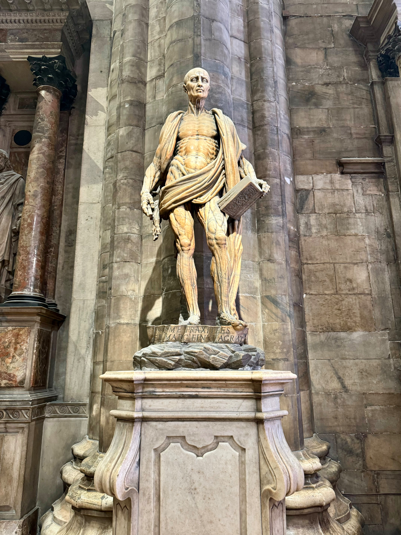 A marble statue of Saint Bartholomew stands in front of a stone wall inside a historical building. The statue depicts an anatomically detailed, muscular figure holding a book and a knife, with a draped cloth over his shoulder.