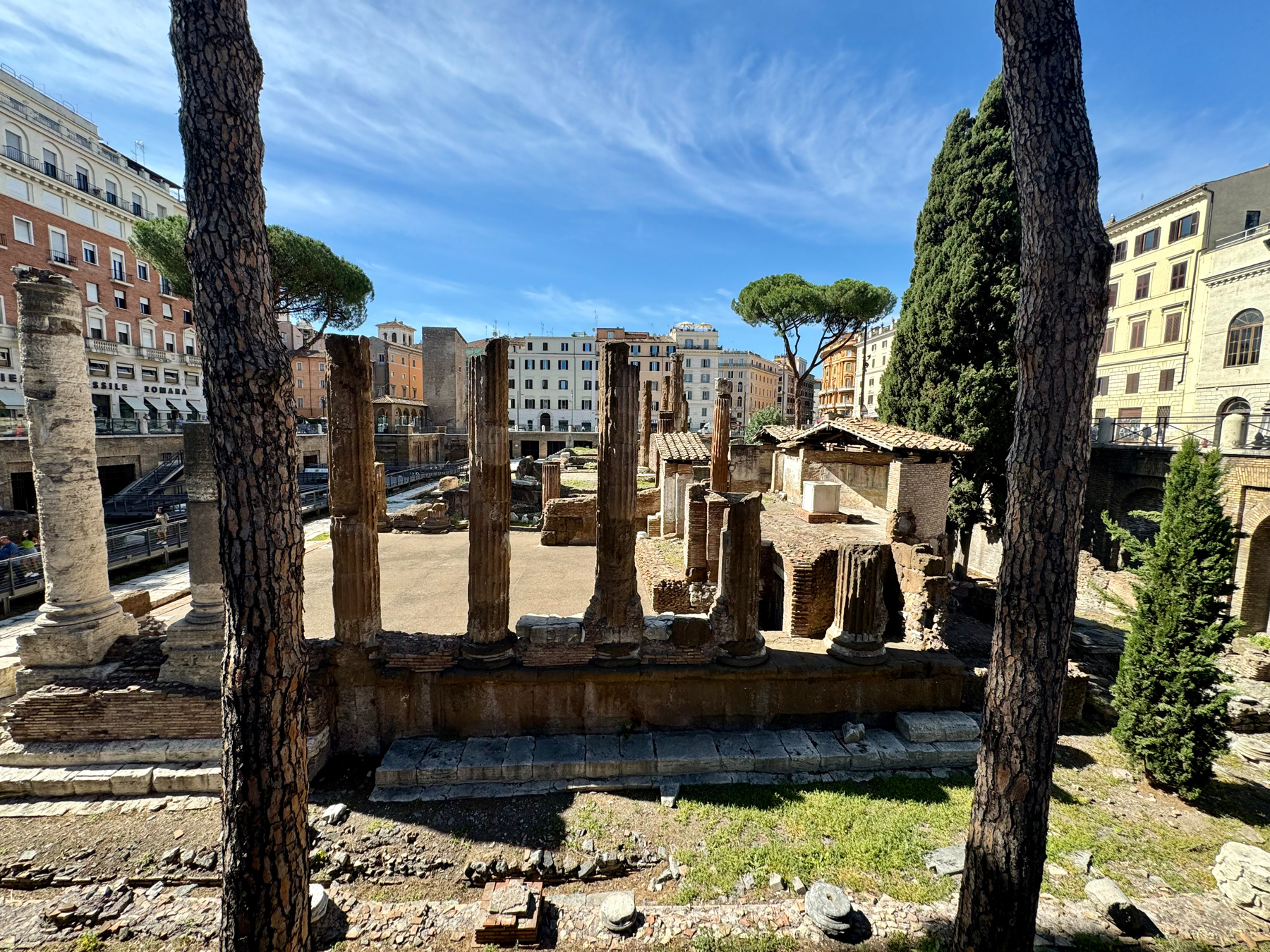 The image depicts the ruins of an ancient Roman temple complex, surrounded by modern buildings, some trees, and columns. The sky is clear and sunny.