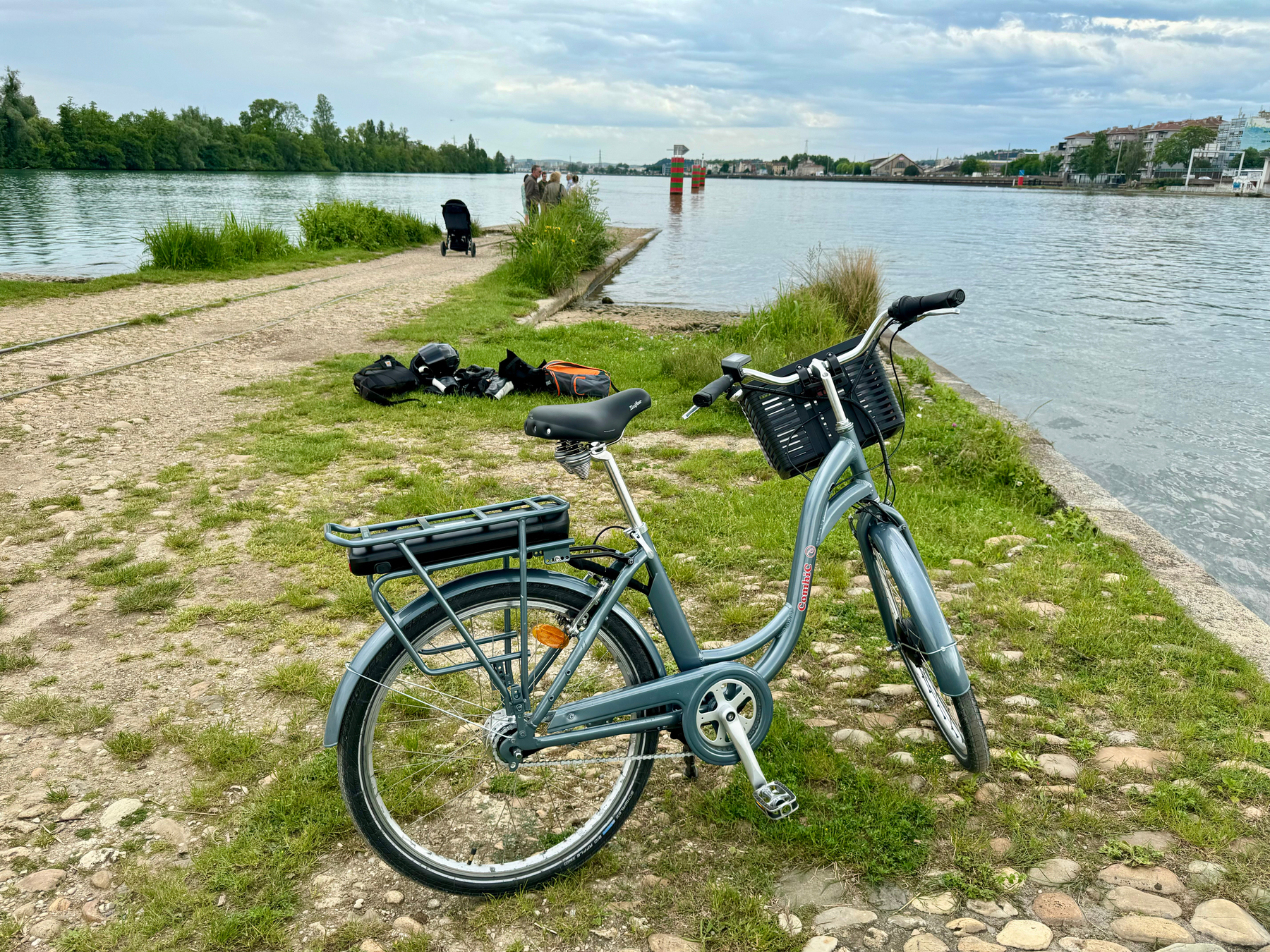 A bicycle parked on a grassy riverbank with personal belongings scattered nearby and people walking in the background. In the background, the confluence of the Saône and Rhône rivers