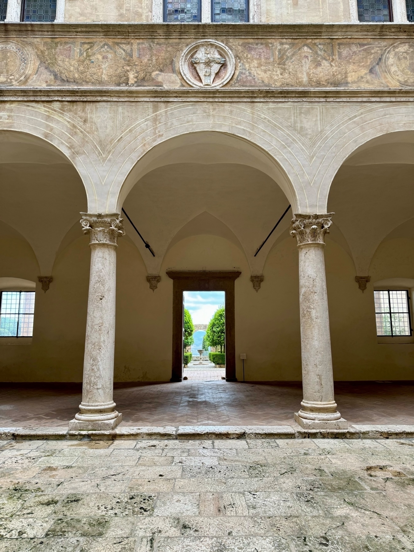 An arched walkway with three columns supporting the structure. Above the arches is a stone wall with faded frescoes and an ornamental carving. Beyond the doorway at the center is a garden pathway lined with hedges leading to a distant view of a lake. 