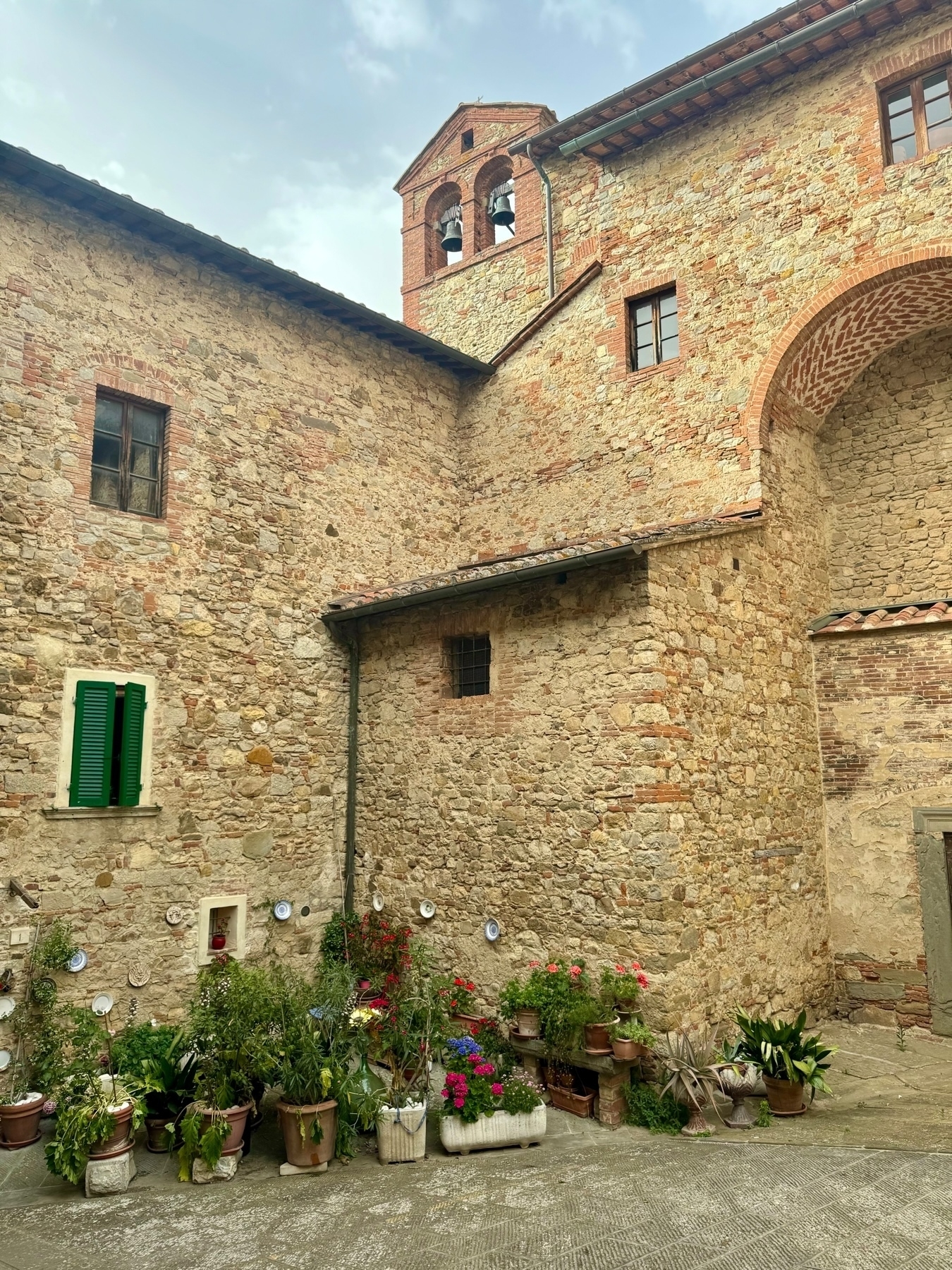 A rustic stone courtyard with potted plants and vibrant flowers arranged against the walls. Decorative plates are mounted on the walls. Above, a bell tower with two bells is visible, and the weathering on the stone walls suggests the structure is historic. 