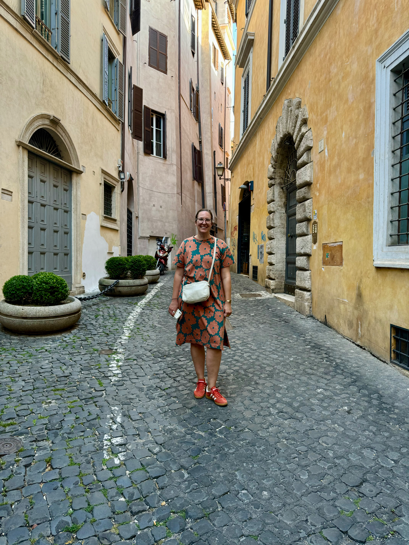 A person standing on a cobblestone street in Rome, surrounded by narrow buildings with wooden shutters and ornate doorways. The individual is wearing a patterned dress and red shoes, and is holding a phone, with a crossbody bag.