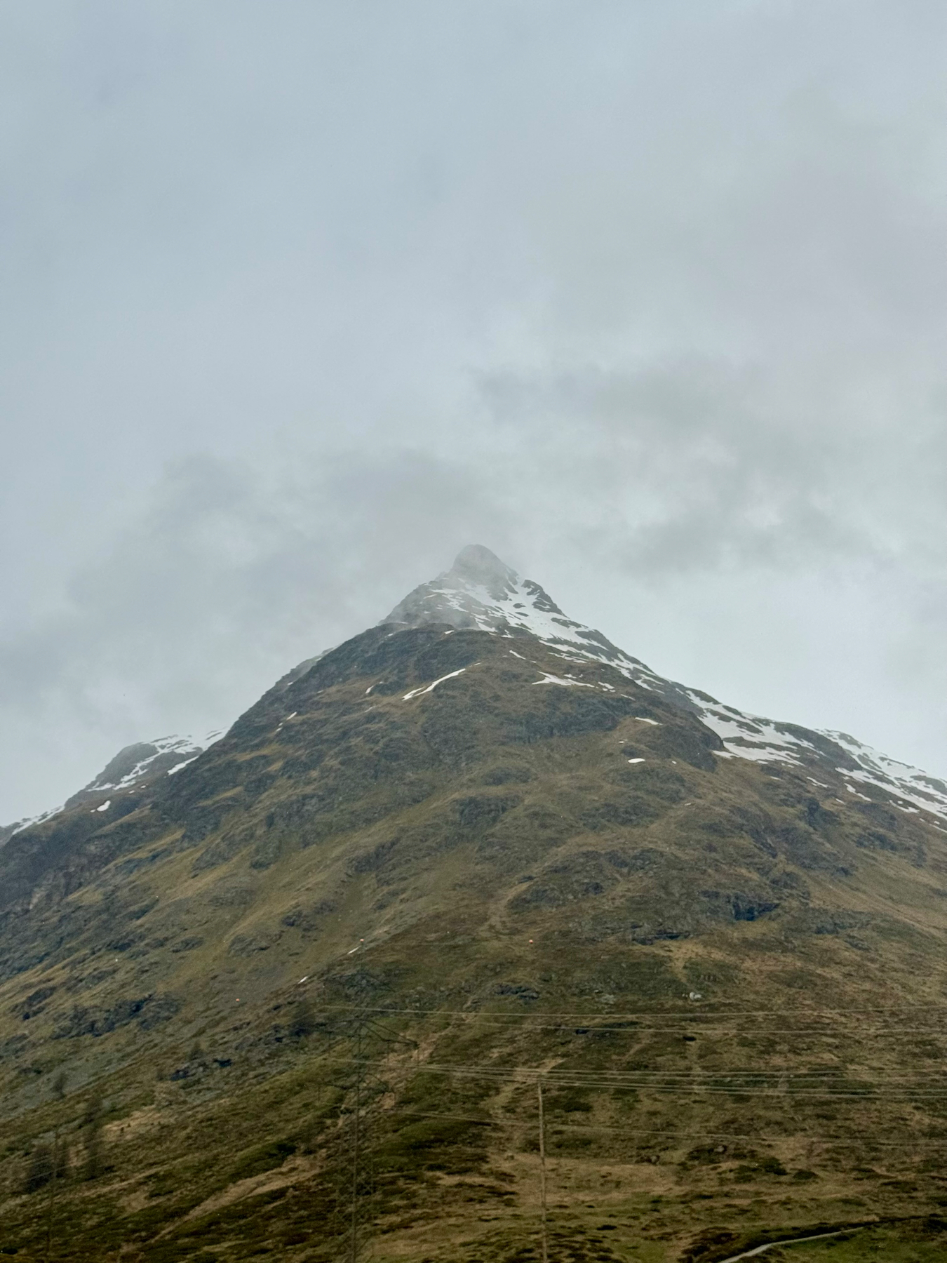 A mountain peak with some snow, against a cloudy sky, with the lower slopes covered in grass and some power lines visible in the foreground.