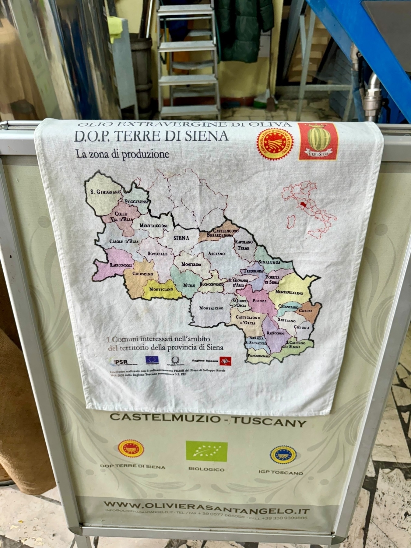 A detailed cloth map depicts the production area for D.O.P. Terre di Siena olive oil in the province of Siena, Tuscany. It highlights various localities involved in olive oil production. 
