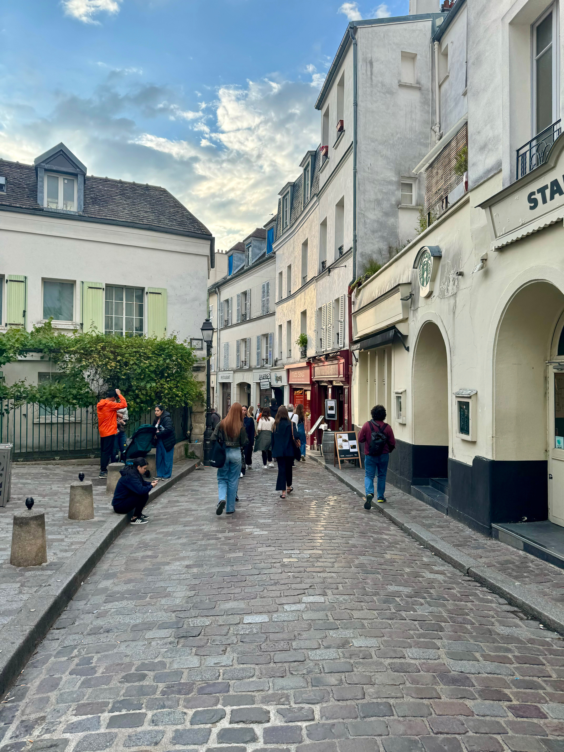 A cobblestone street in a quaint city quarter with pedestrians walking and some people taking photographs. Classic European buildings line the street, and there is a “Starbucks Coffee” sign on the right side.