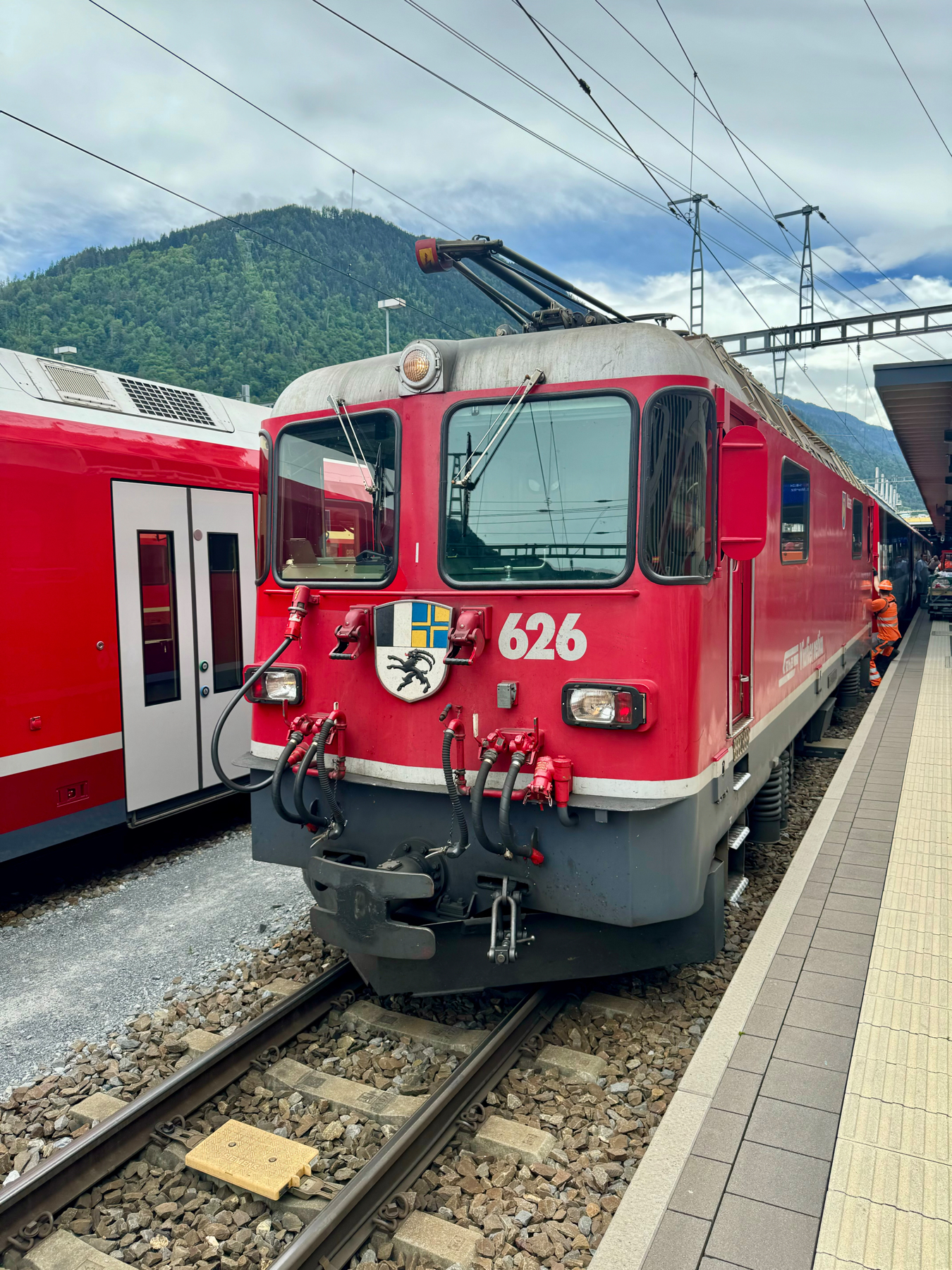 A red train locomotive with the number 626 parked at a train station, with mountains and a cloudy sky in the background.