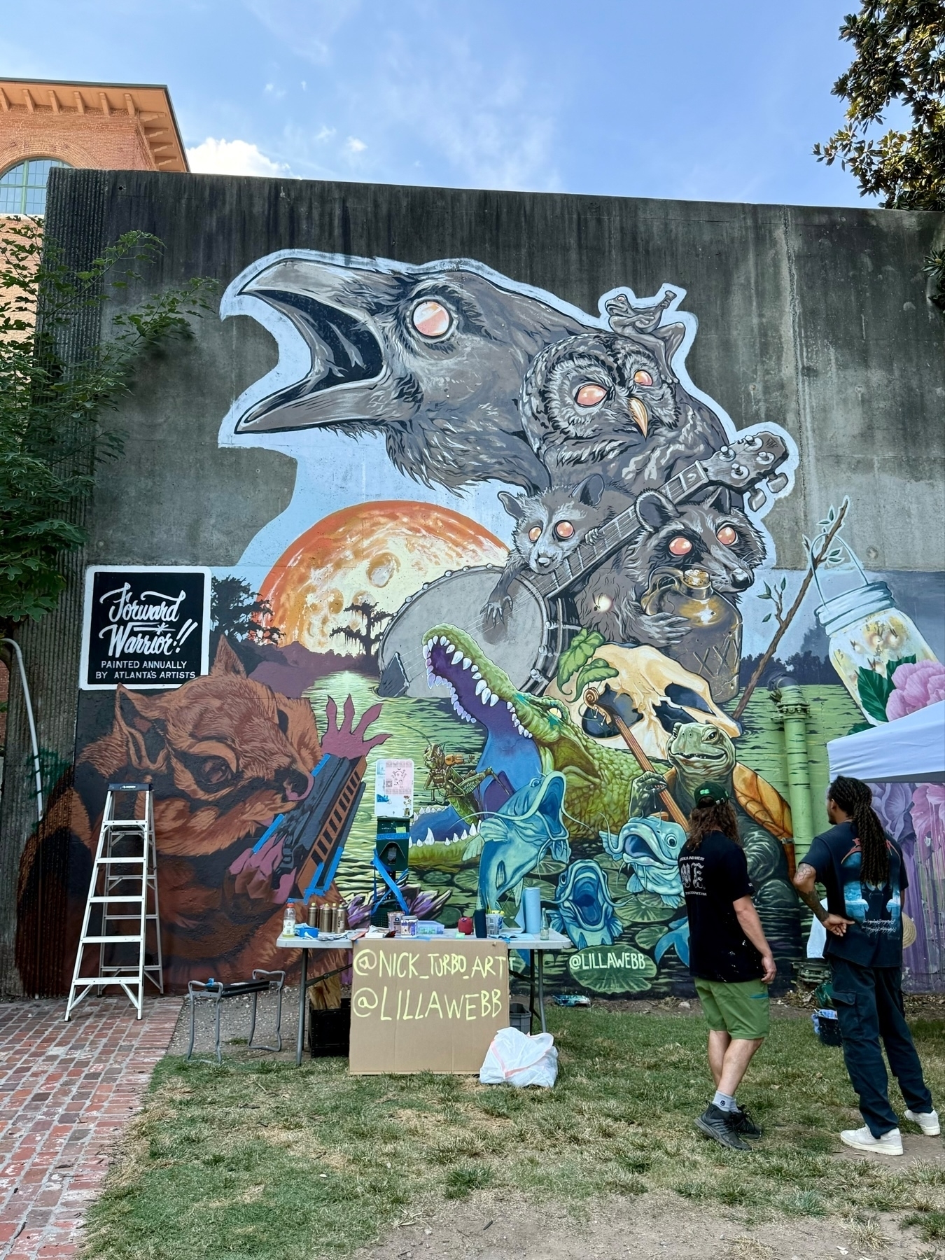 A large, colorful mural on a concrete wall depicting a range of fantastical creatures, including a large crow, an owl, various small animals, and a green alligator-like creature holding instruments. Two people stand in front of the mural, observing it. 