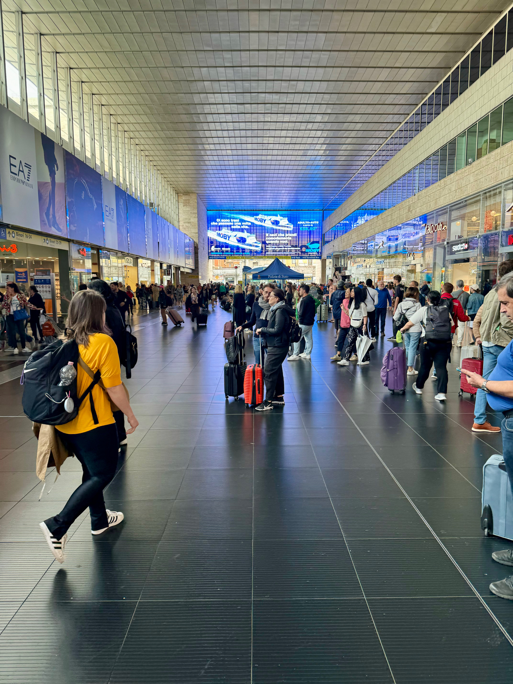 A busy interior of a train station with travelers pulling suitcases and standing in line. The station features a high ceiling, large digital displays, and various shops along both sides. The floor is tiled in black.