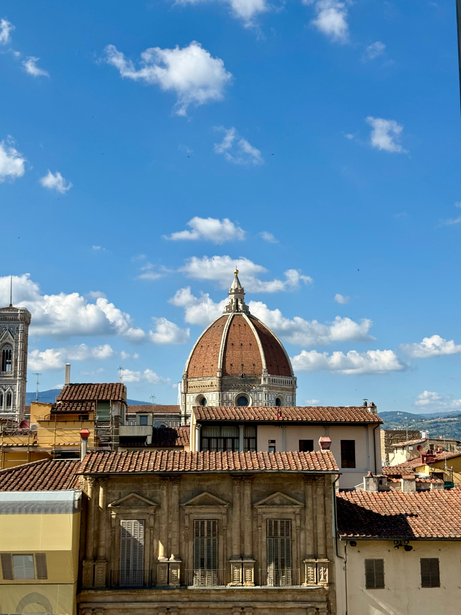 A cityscape featuring the dome of the Florence Cathedral (Duomo di Firenze) in the center, with surrounding historic buildings and rooftops. The sky above is blue with scattered clouds.