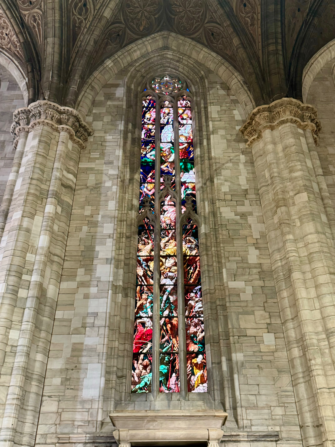 A tall, narrow stained glass window set in a gothic-style arched frame within a stone cathedral wall. The window features intricate depictions of religious scenes and figures in vivid colors. The surrounding architecture includes ornate stone columns and a vaulted ceiling.