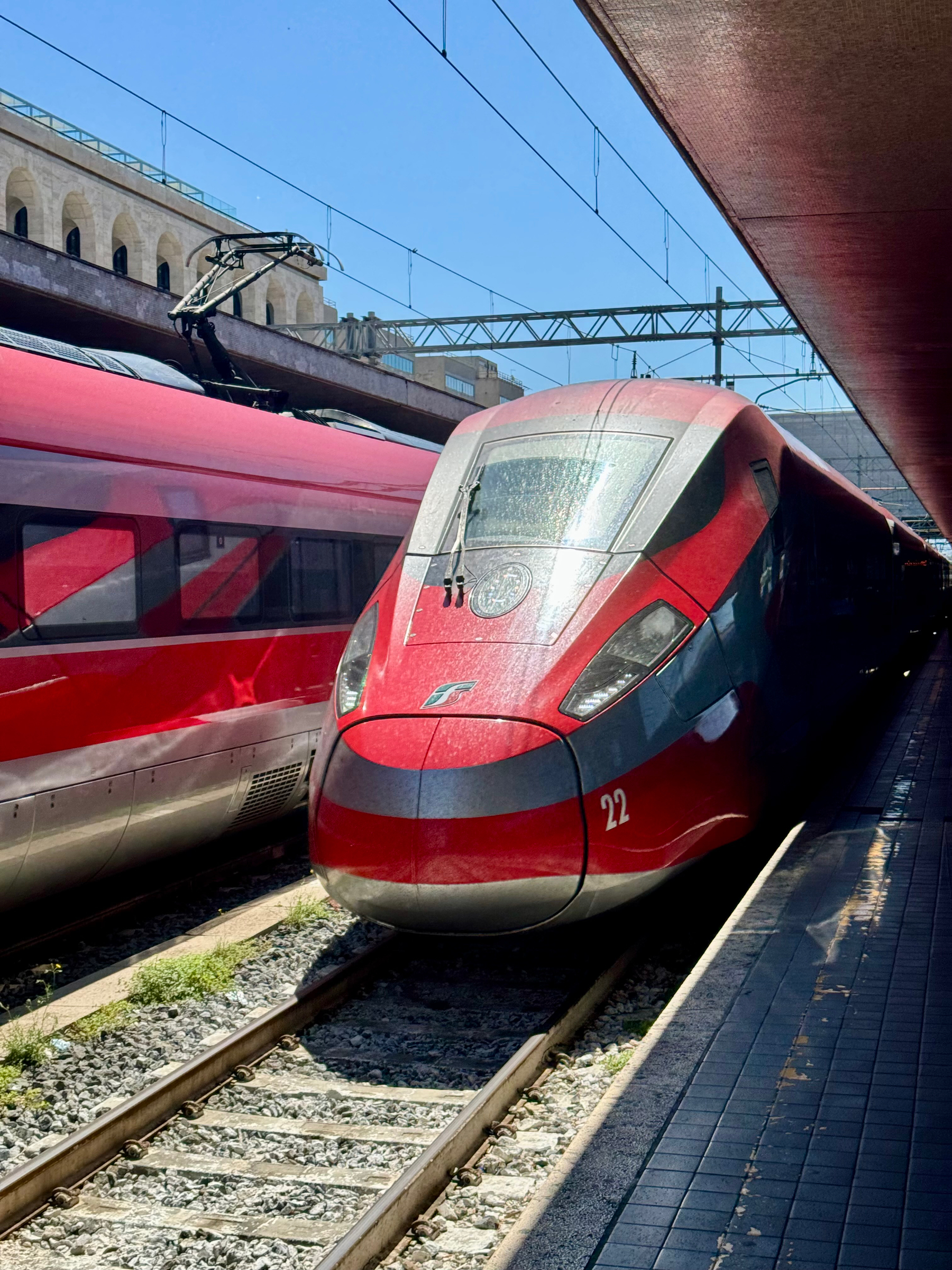 A modern high-speed train at a station platform under a clear blue sky. The train has a red and silver design, with the number “22” visible on its front. Another similar train is on an adjacent track. 