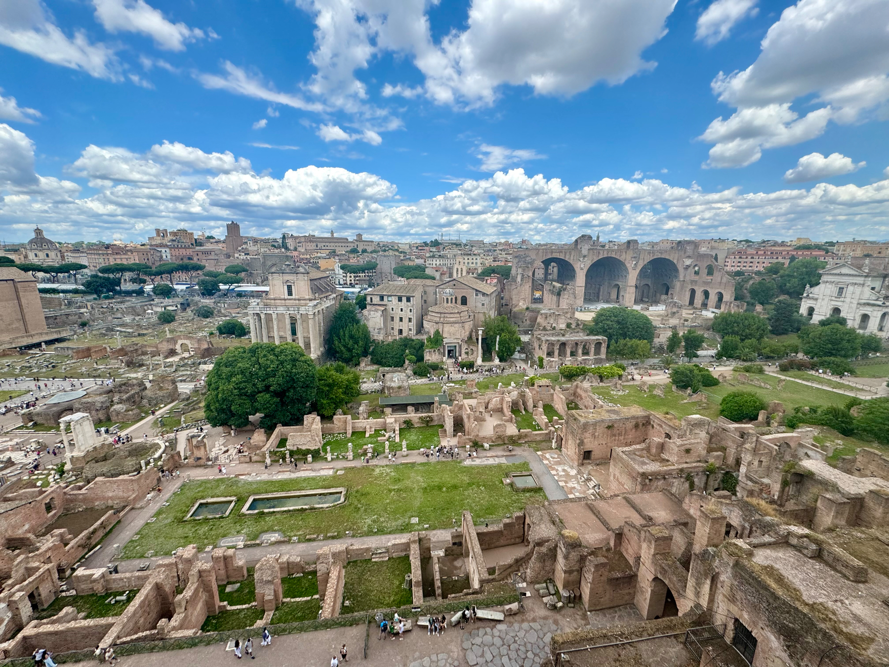 Panoramic view of the Roman Forum in Rome, Italy, showcasing ancient ruins including temples, columns, and stone structures. The vast archaeological site is dotted with greenery and visitors exploring. The background features more historic buildings and arches set against a blue sky.