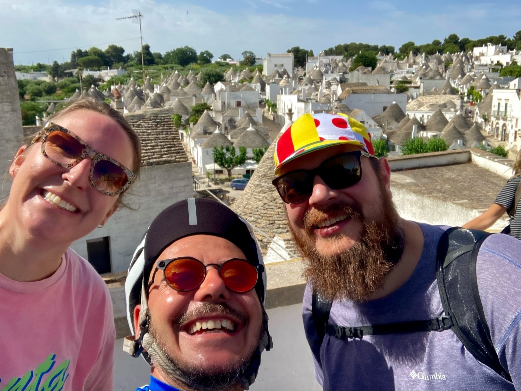 Three smiling individuals are taking a selfie in front of a scenic background featuring traditional white conical-roofed houses. One person is wearing a bike helmet, another has a colorful cap, and the third is wearing sunglasses. The background shows a picturesque landscape. 