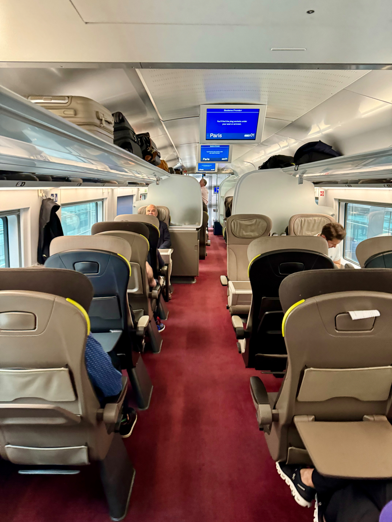 Interior of the Eurostar high-speed train car with business class seating. The seats are arranged in pairs, upholstered in grey with yellow accents. Overhead luggage racks hold various bags and suitcases. Digital screens display information about the destination, which is Paris. Pass