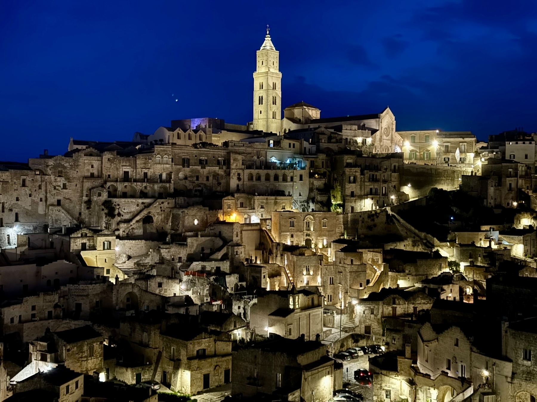 View of Matera, Italy at night with the illuminated historic Sassi (stone houses) and the Cathedral of Matera prominently visible against a dark blue sky.