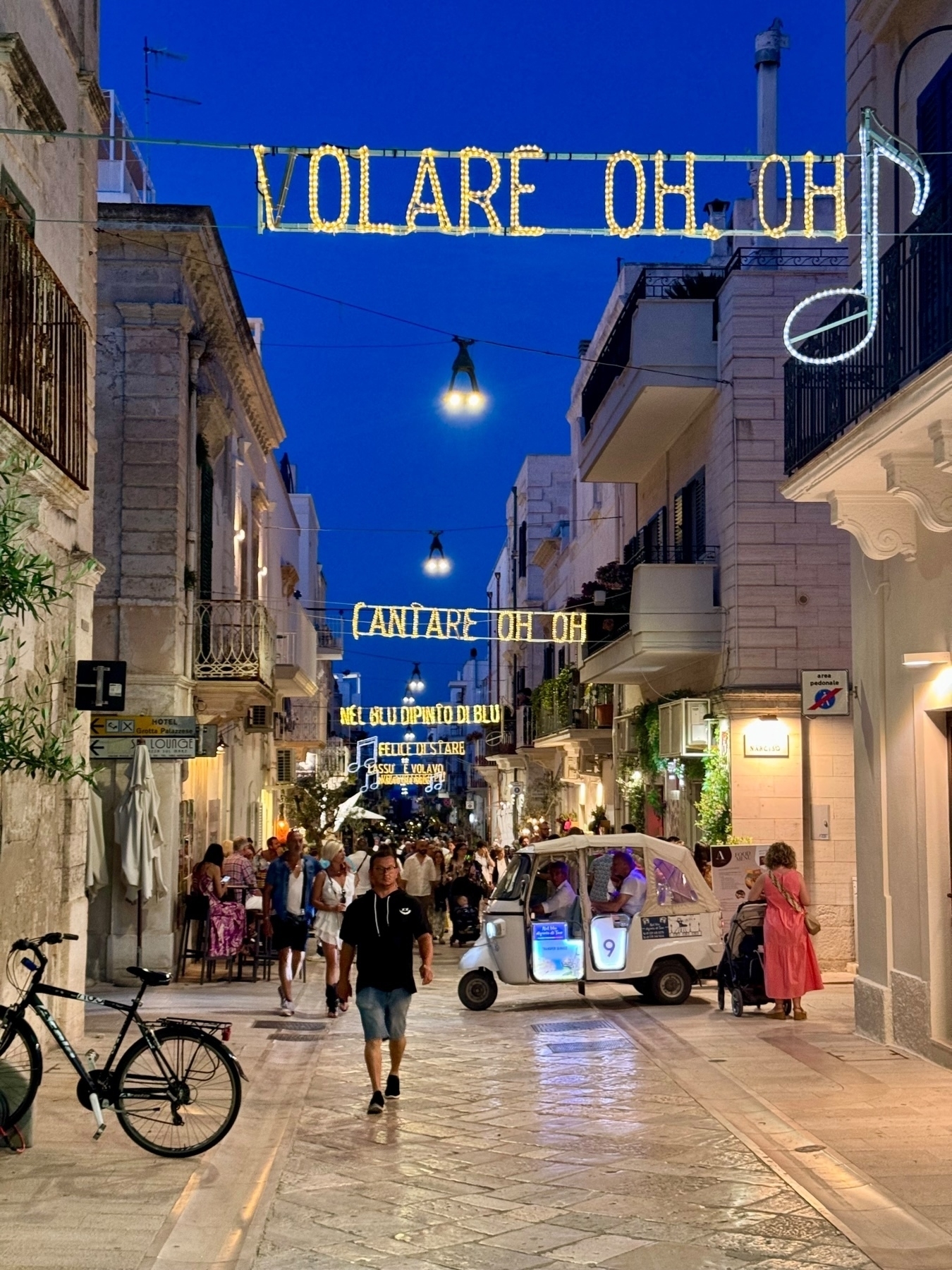 A charming evening street scene in an Italian town. Decorative lights hang above, displaying the words “VOLARE OH OH” and a musical note. The street is bustling with people walking and enjoying the evening.