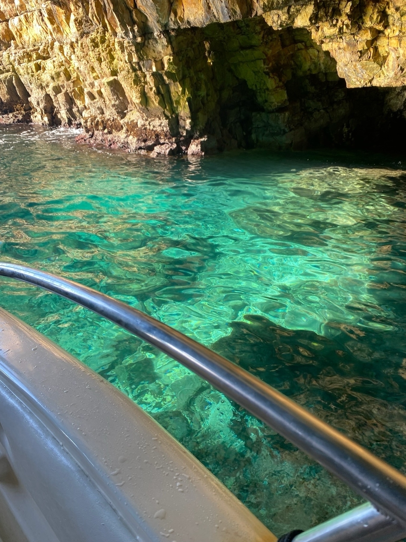 A small boat navigating close to a rocky cliff with a shallow cave. The water is clear and turquoise, reflecting the surrounding rocks. The boat’s railing and part of the hull are visible in the foreground.