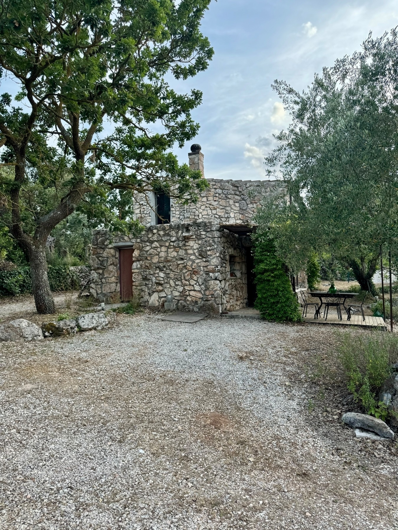 A rustic stone house surrounded by trees and greenery. There is a small patio area with a metal table and chairs on the right. The ground is covered in gravel and there is an old wooden door on the house. The sky is partly cloudy.