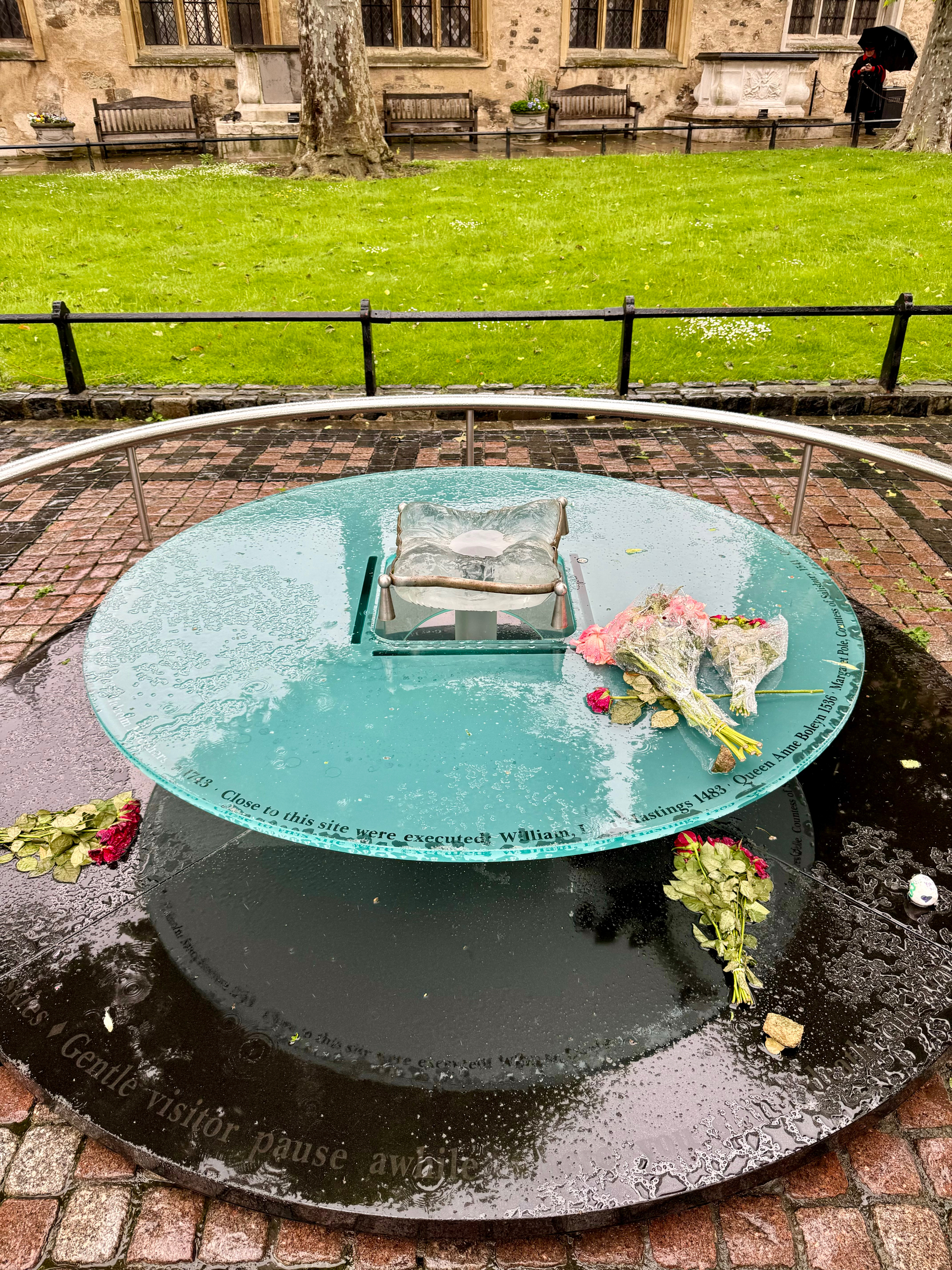 A glass memorial plaque on wet ground surrounded by a metal railing with raindrops and flowers laid on top, inscribed with text commemorating the execution site of historical figures, including Anne Boleyn. 