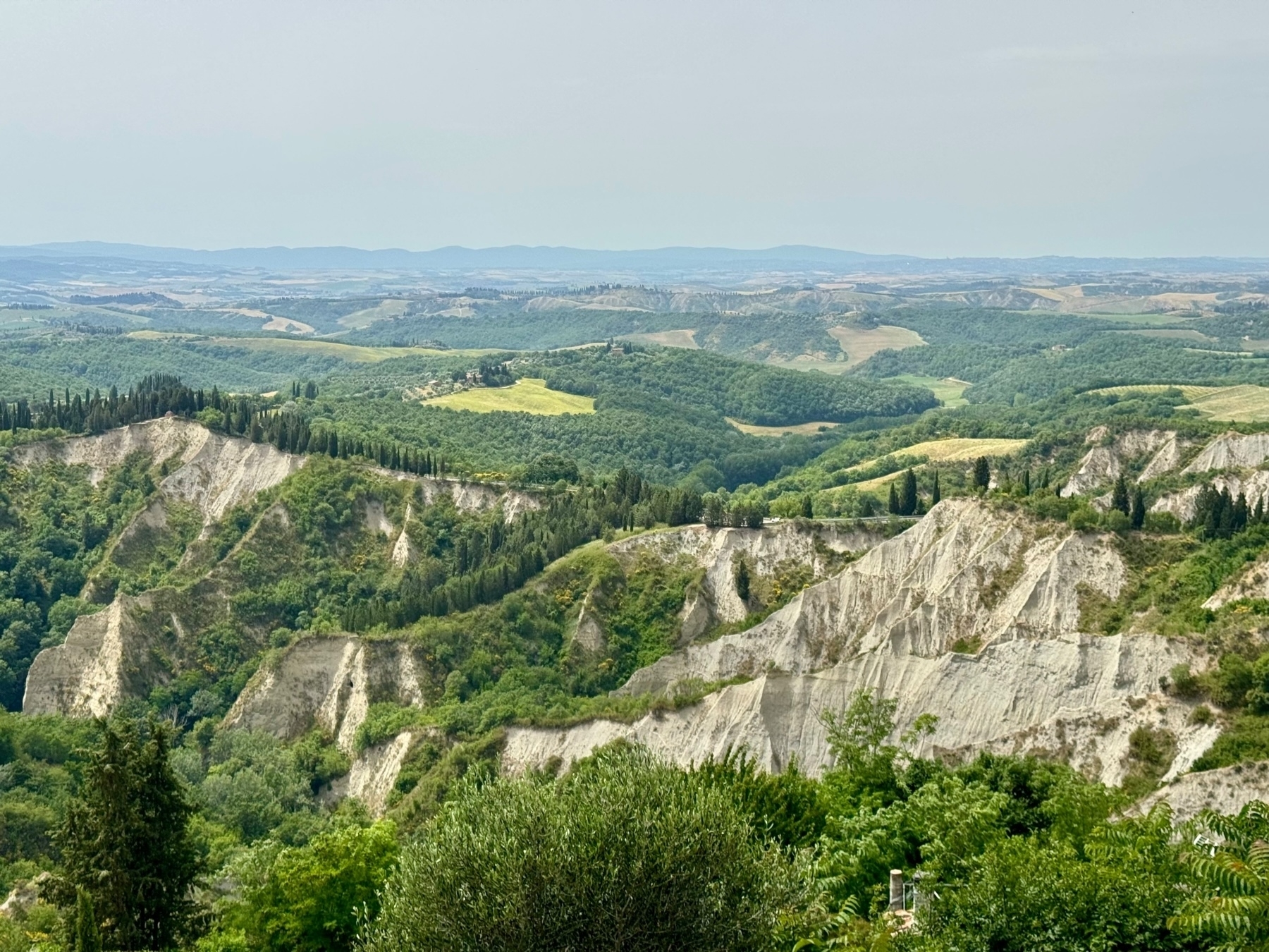 A panoramic view of a hilly landscape featuring rugged, white chalk formations and lush green vegetation. Rolling hills and dense forests stretch into the distance, with patches of farmland visible under a clear, light blue sky.