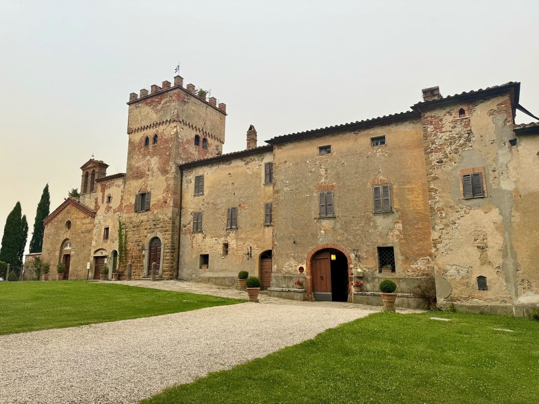 A historic, weathered stone building with a tall, crenellated tower, arched doorways, and shuttered windows. The structure includes both medieval and Renaissance architectural elements with a large, grassy courtyard and a gravel path leading to the entrance.