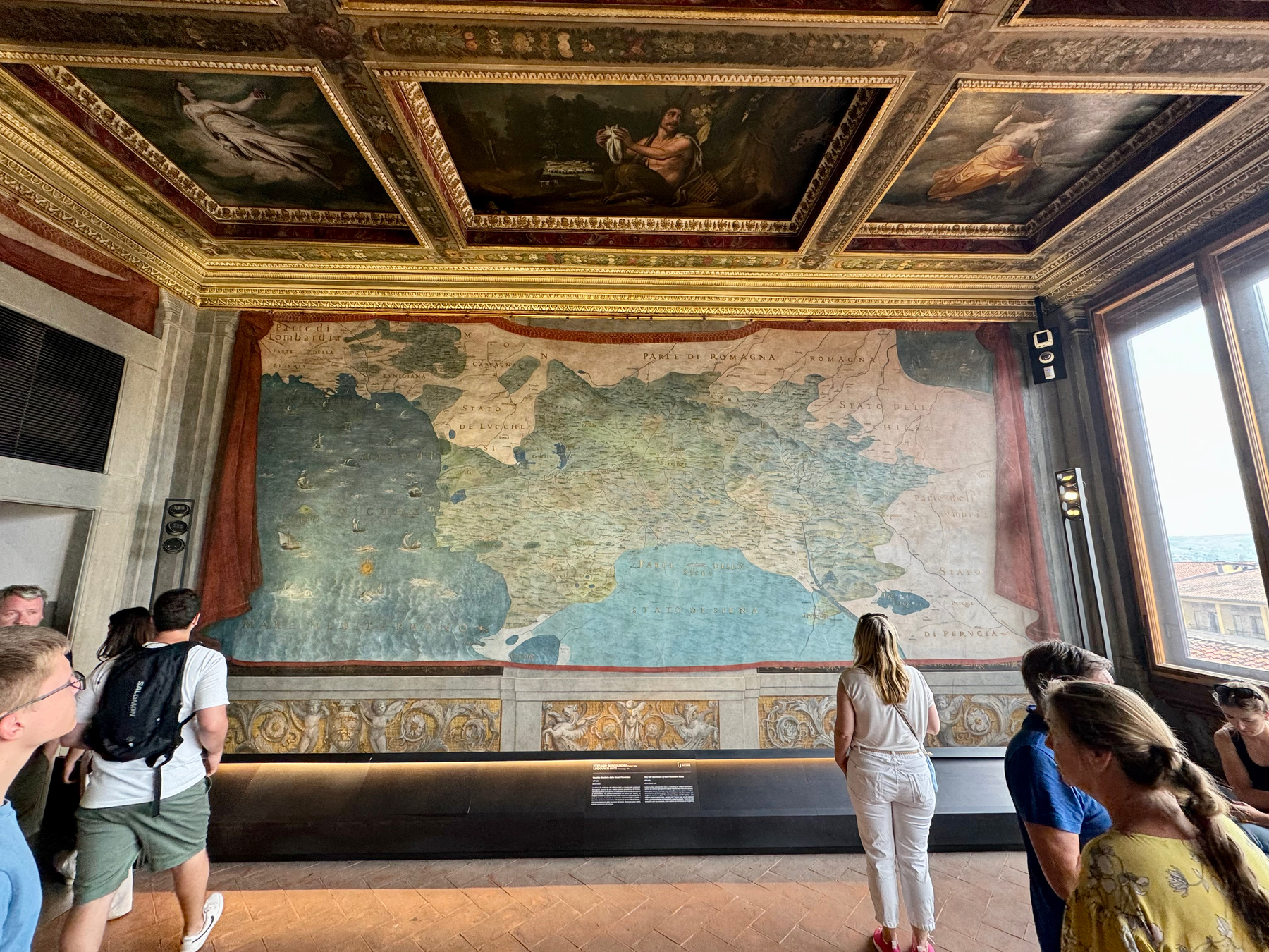 A group of people is viewing a large, historic map of Italy displayed on a wall in a room with ornate gold-trimmed ceilings and detailed paintings. Natural light pours in from a window on the right side of the image.