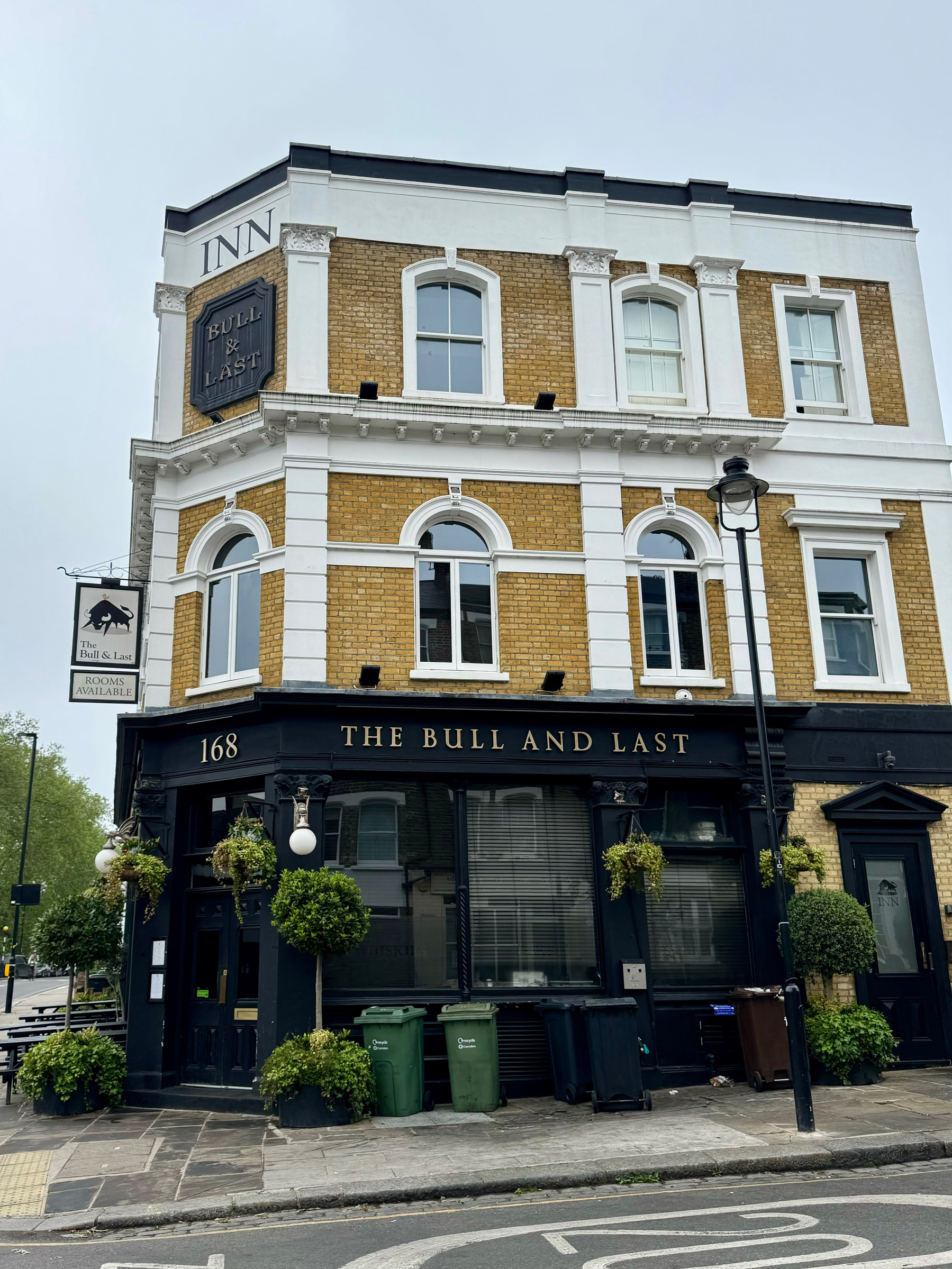 A traditional brick building housing “The Bull and Last” pub with a black facade, large windows, hanging plants, and road signs visible on a cloudy day.