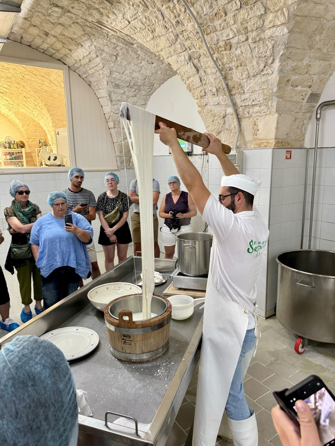 A group of people, wearing hair nets, watches a cheesemaker demonstrating the process of making cheese in a rustic, stone-vaulted room. The cheesemaker is pulling and stretching the cheese curds, which are cascading into a wooden barrel.