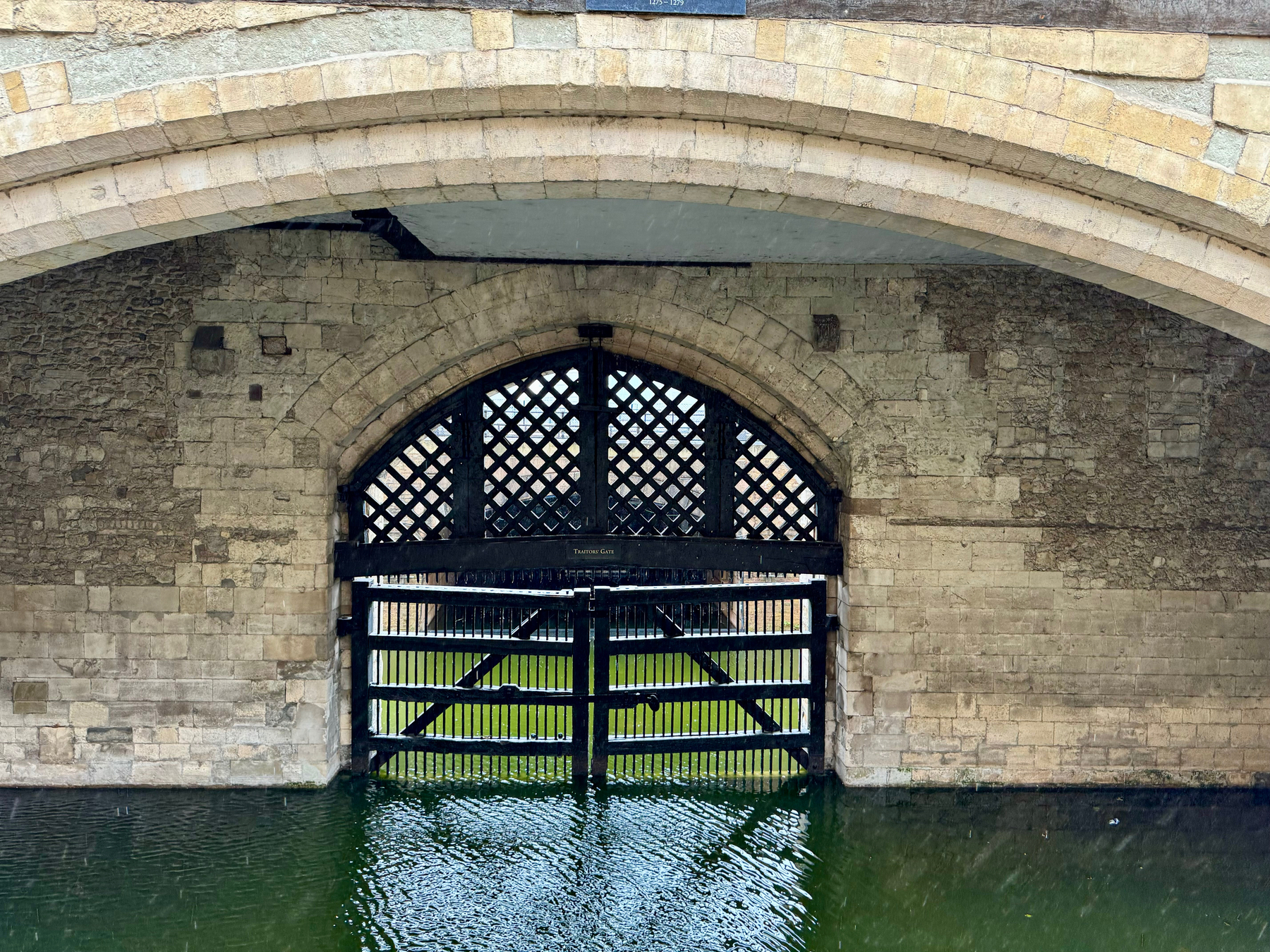 Stone bridge arch with a black lattice gate partially submerged in water, reflecting in the water below. A plaque reads “Traitors' Gate.