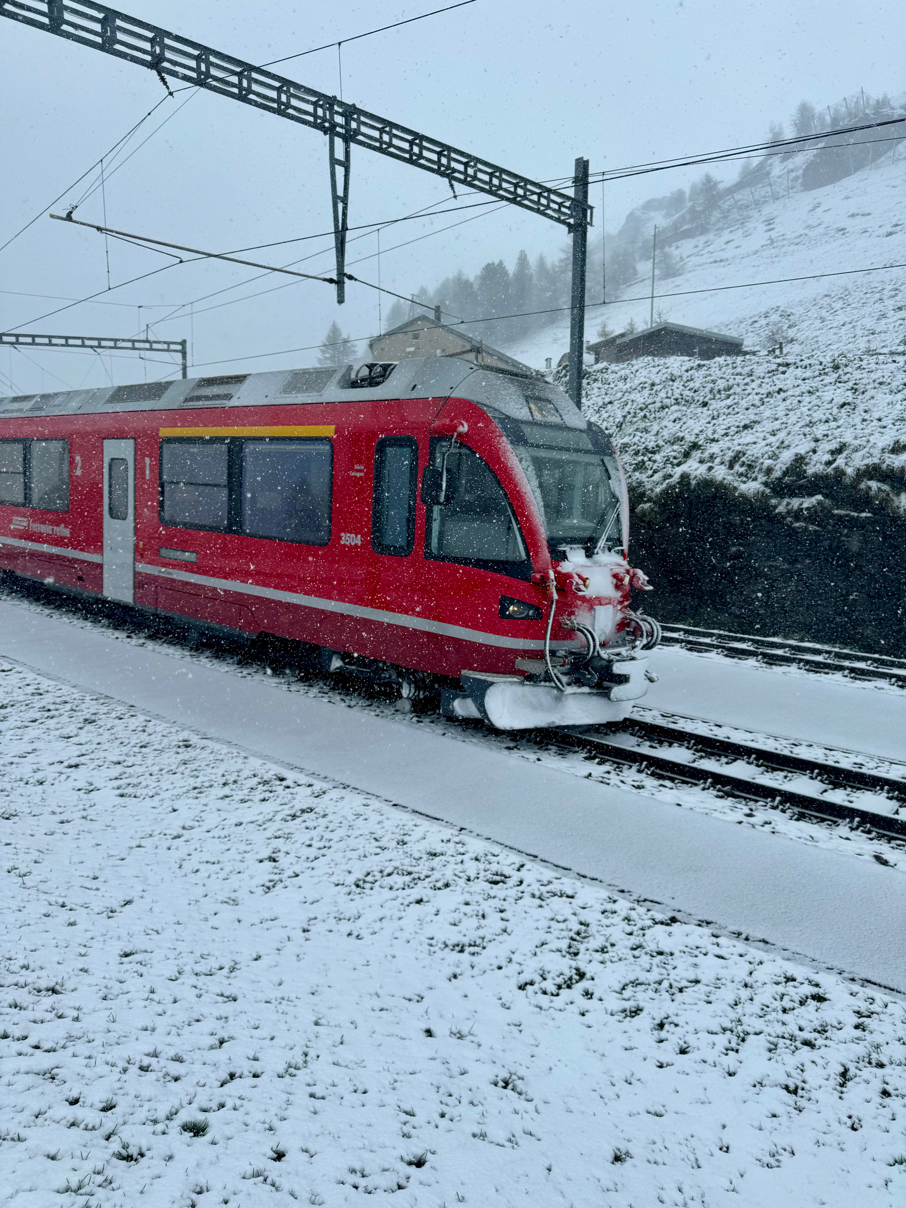 A red train is traveling through a snowy landscape. The tracks and the surrounding area are covered with snow. Overhead, electric lines span above the train, and a hill with some buildings is visible in the background under a light snowfall.