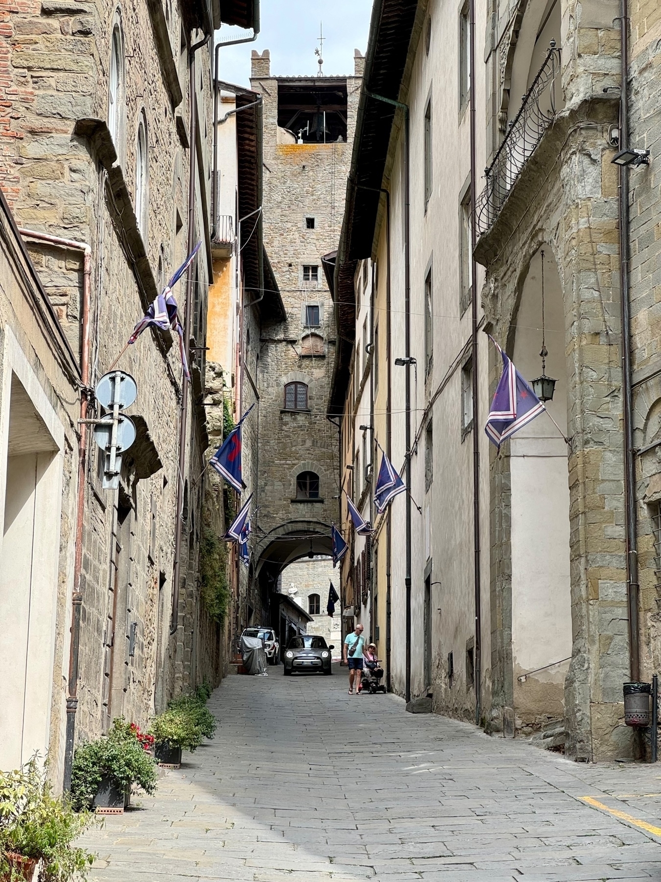 Narrow street in a historic European town, lined with stone buildings and colorful banners. A steep incline leads to an archway and a tower in the background. A few pedestrians and a parked car are visible.