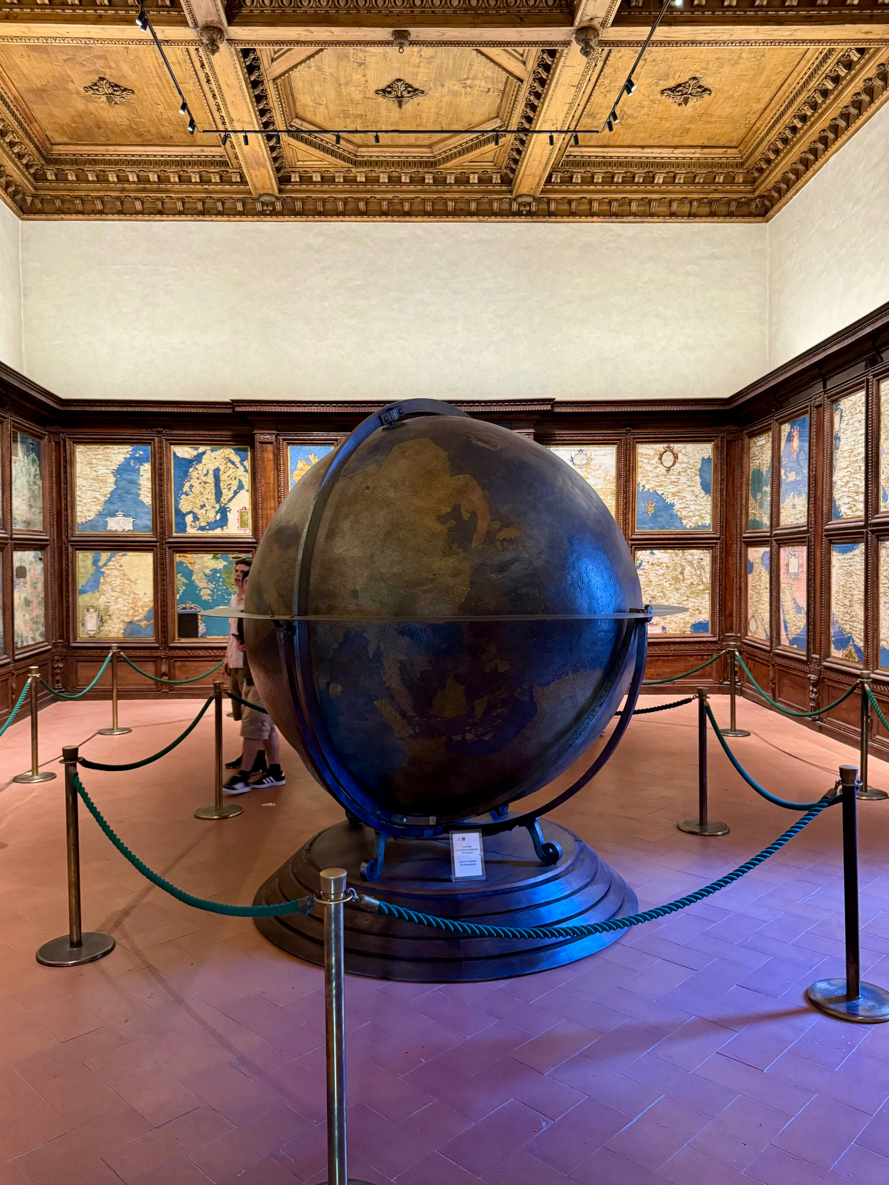 A large, antique globe is displayed in the center of a room surrounded by wooden paneling and world maps. The ceiling is ornately decorated, and the globe is enclosed by ropes to keep viewers at a distance. 