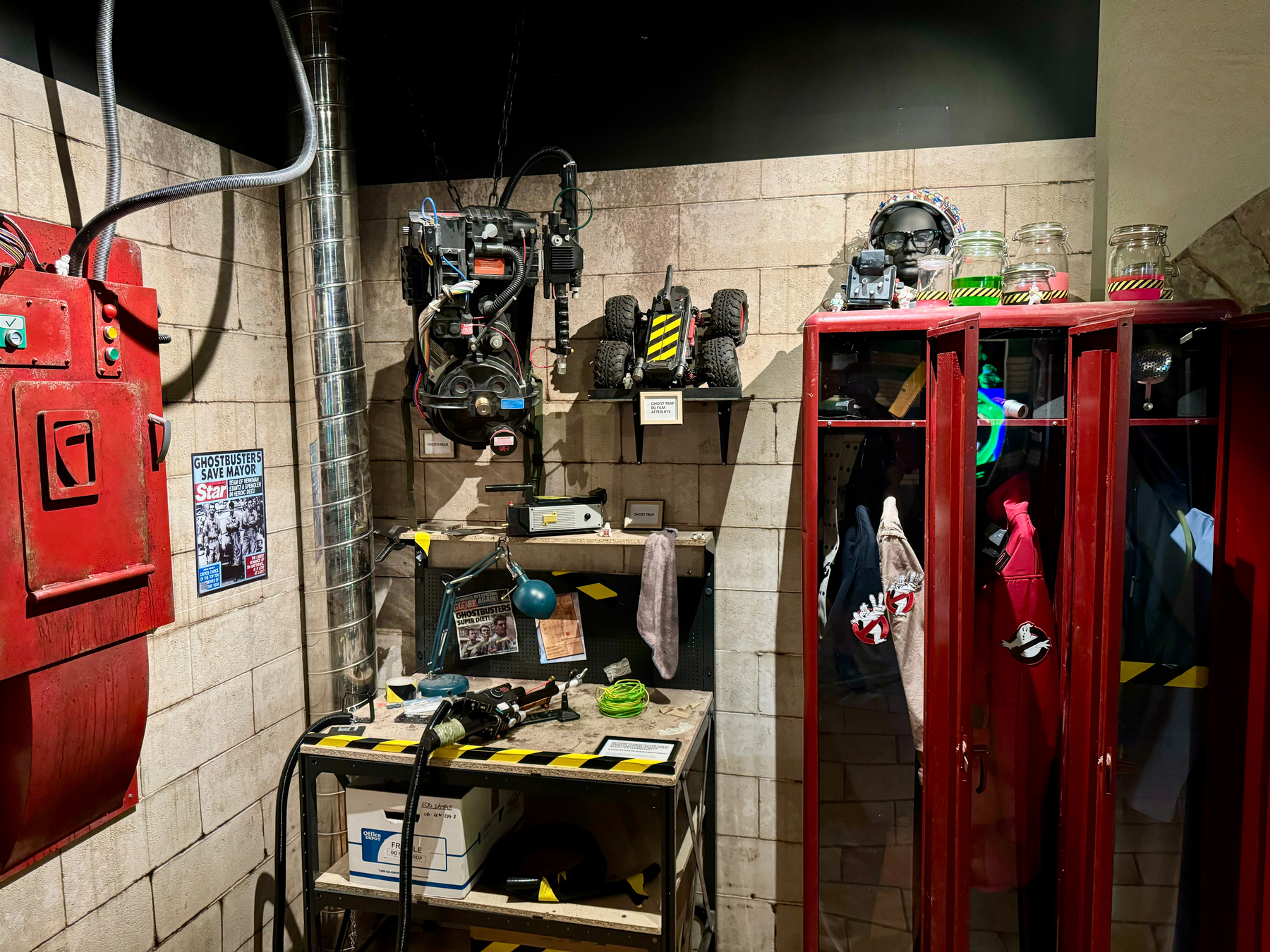 A Ghostbusters-themed display featuring a Proton Pack, ghost traps, a PKE Meter, lockers with jumpsuits, and themed collectibles.