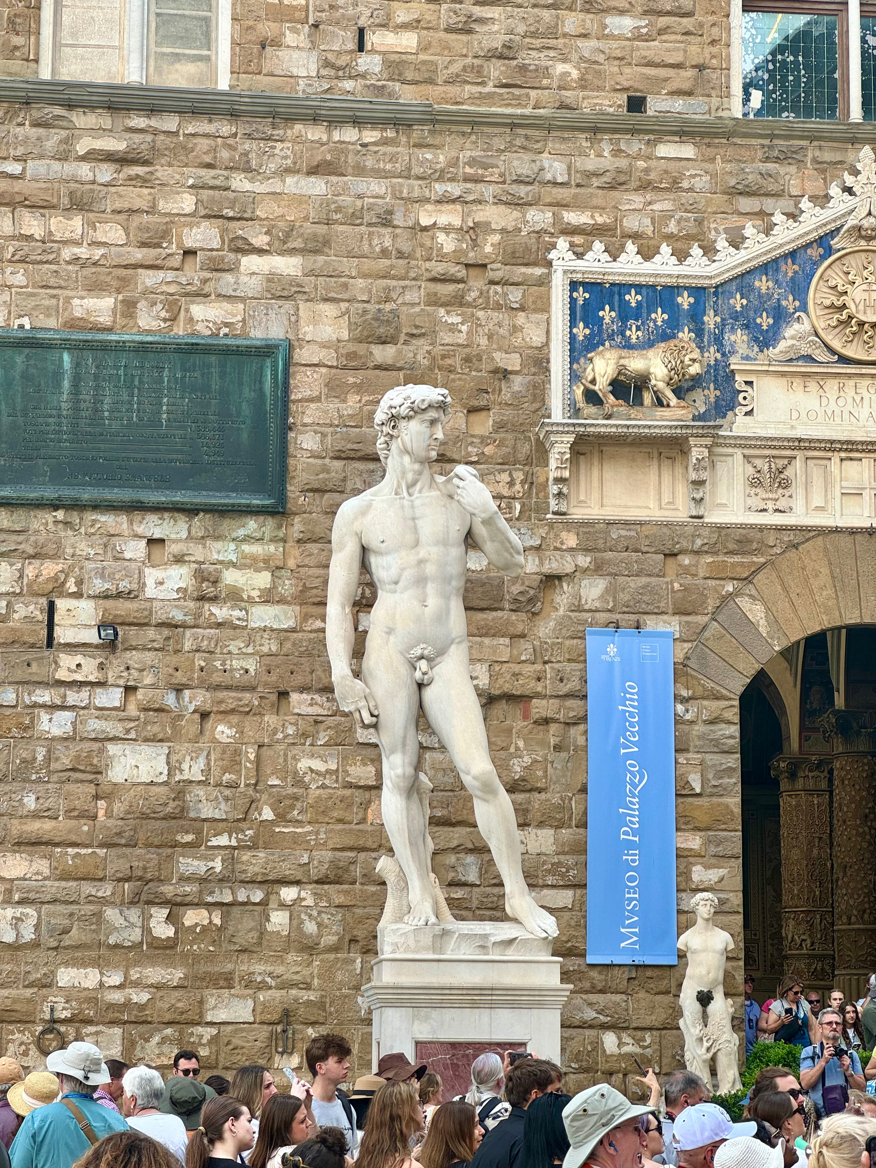 The image captures a replica of Michelangelo’s statue of David displayed outside the Palazzo Vecchio in Florence, Italy. Surrounding the statue is a crowd of tourists. The stone facade of the Palazzo Vecchio and a blue banner for the museum are visible. 