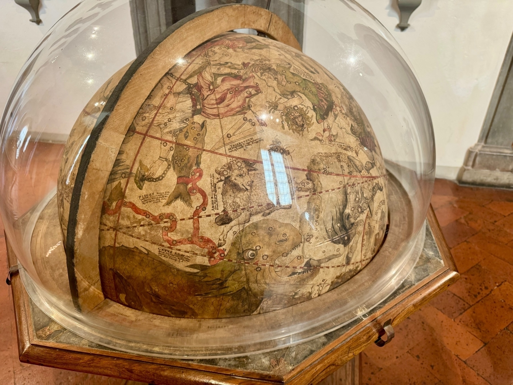A historical celestial globe encased in a glass dome. The globe features detailed illustrations of constellations with mythological figures and celestial coordinates. The setup is placed on a wooden stand, and its surroundings indicate an indoor museum or exhibit space.