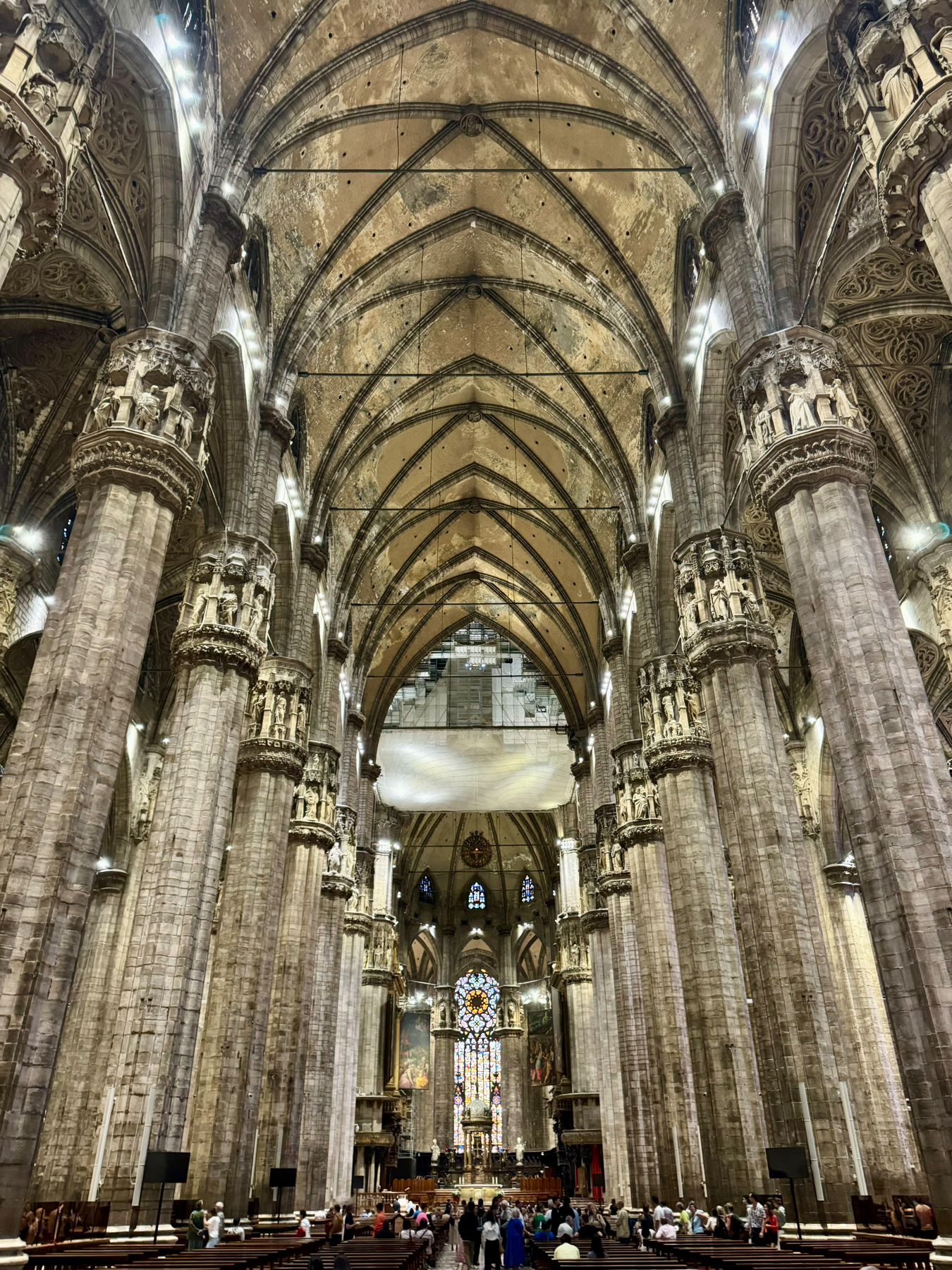 Interior of a grand Gothic cathedral with towering columns, arched ceilings, ornate architectural details, stained glass windows, and people seated in pews and walking around.