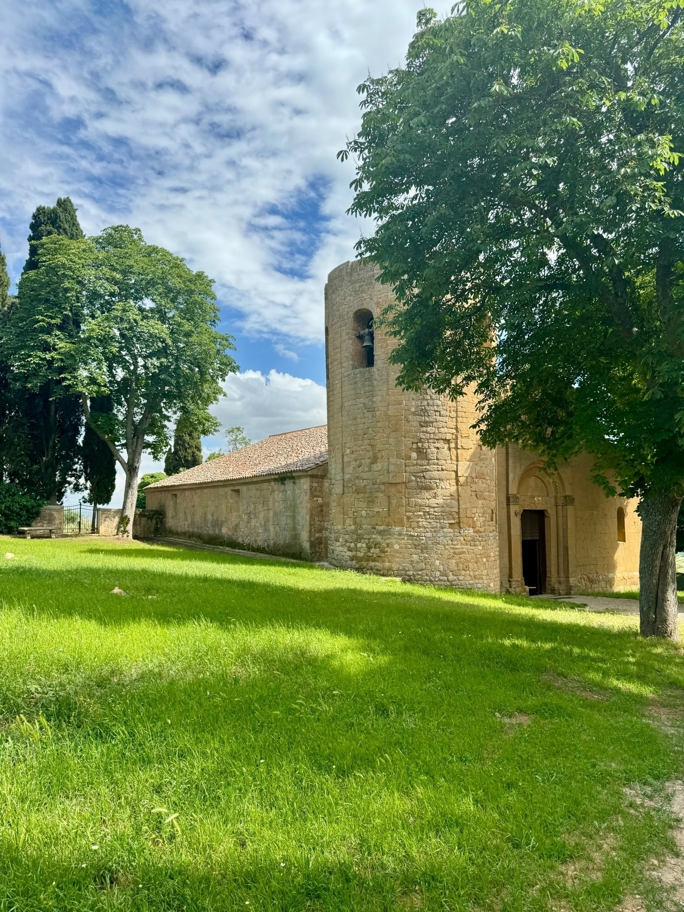 A historic stone church with a cylindrical tower and arched entrance, surrounded by lush green grass and leafy trees under a partly cloudy sky.