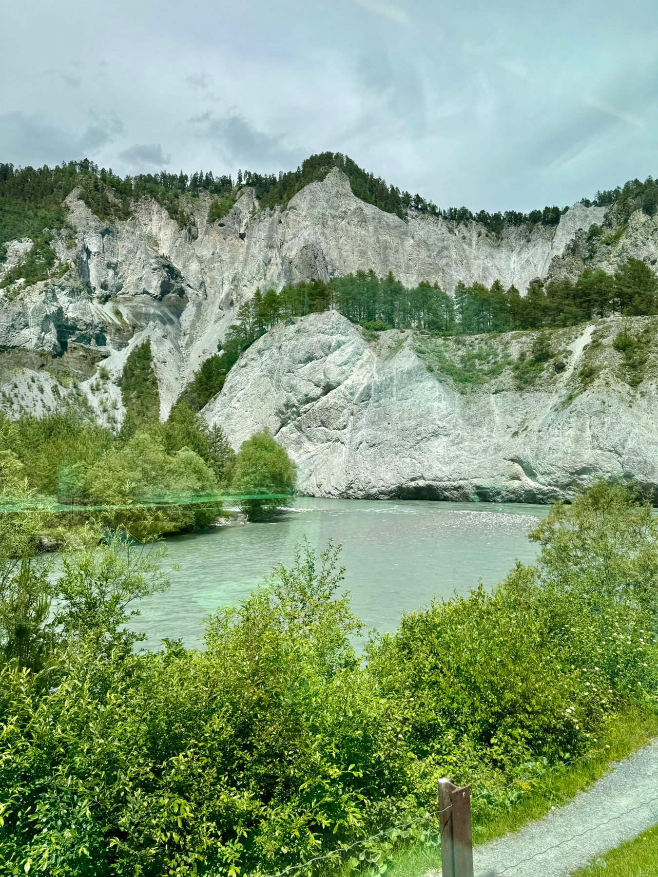 A serene landscape featuring a turquoise river in the foreground with lush greenery on its banks and dramatic, rocky mountains in the background, all under a slightly cloudy sky.