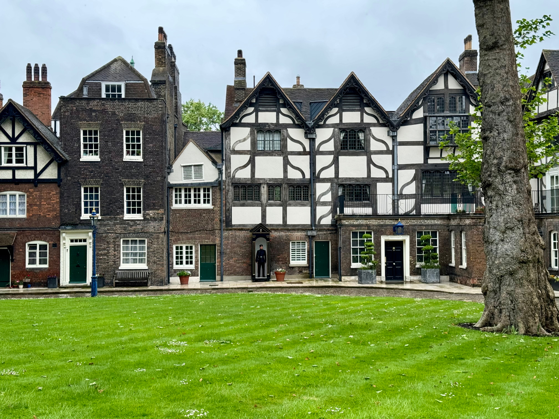 The Queen’s house among other traditional English Tudor-style buildings with white and black timber framing, surrounding a green lawn with a tree to the right. There is a person standing by the entrance of the central building.