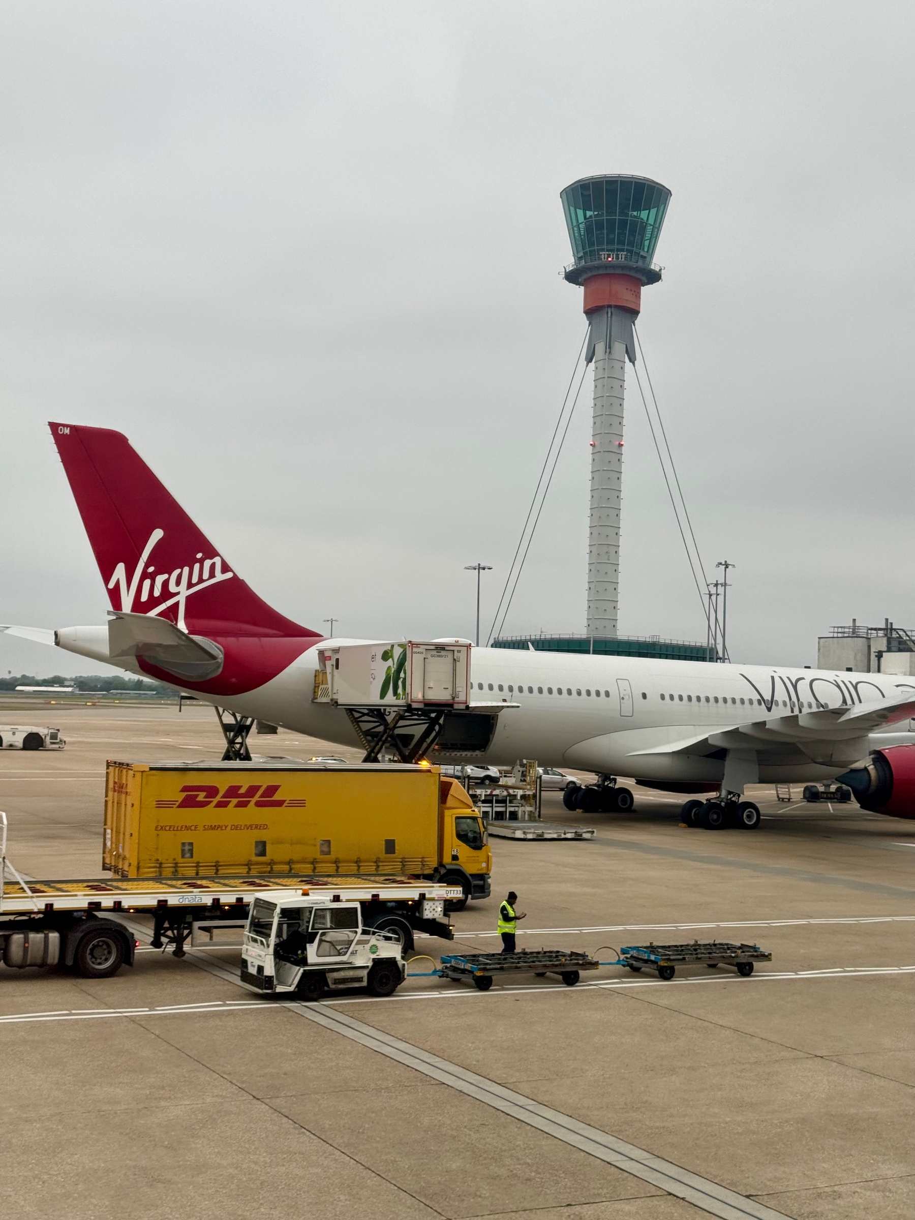 An airport apron with a Virgin airline aircraft, a DHL delivery truck, a baggage tug, an air traffic control tower, and a ground crew member wearing a hi-vis vest.
