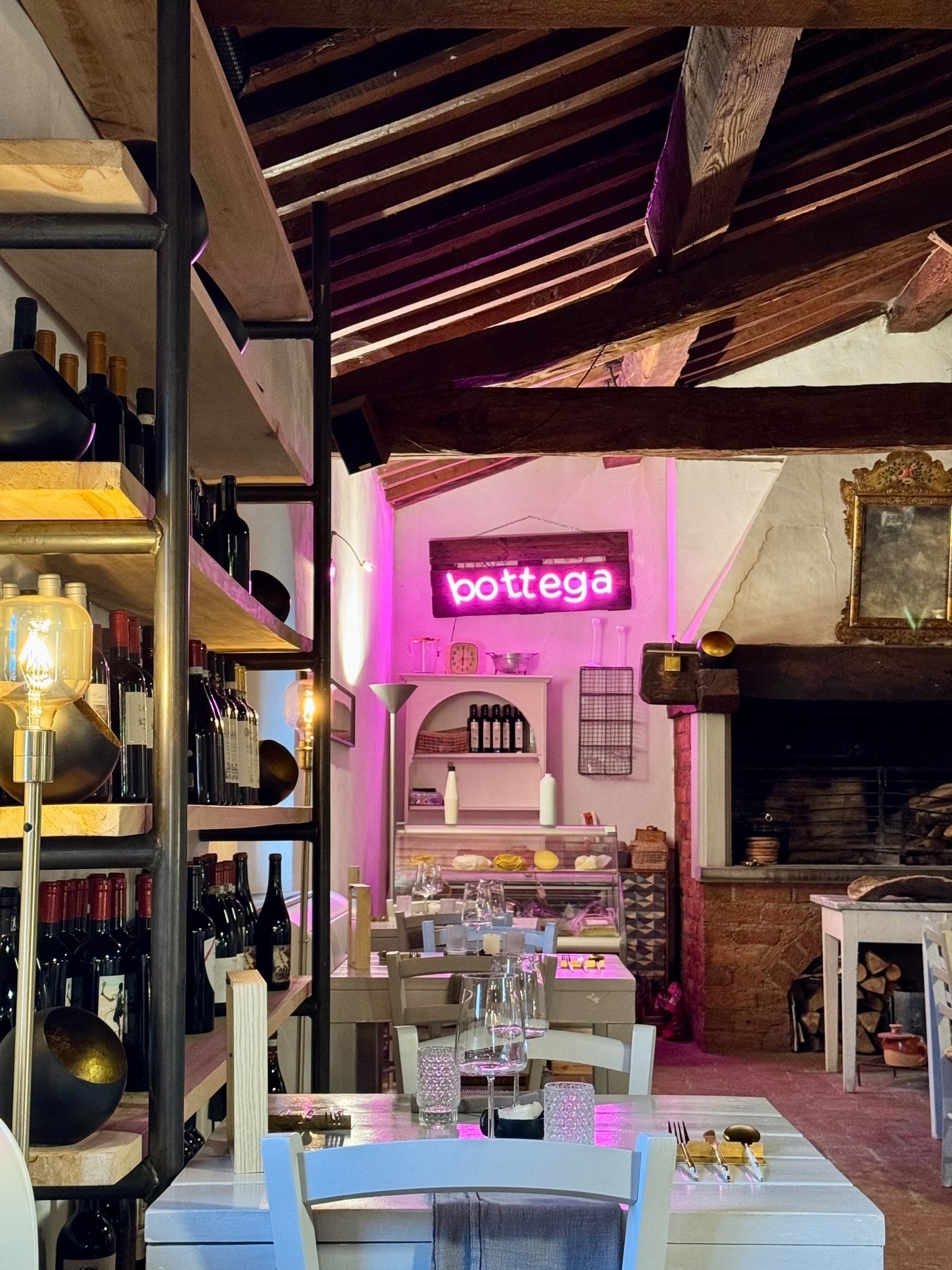 A cozy dining area with rustic wooden beams and shelves filled with wine bottles. The tables are set with glasses, cutlery, and candles. A neon sign reading “bottega” is illuminated in pink on the back wall, adding a modern touch. 